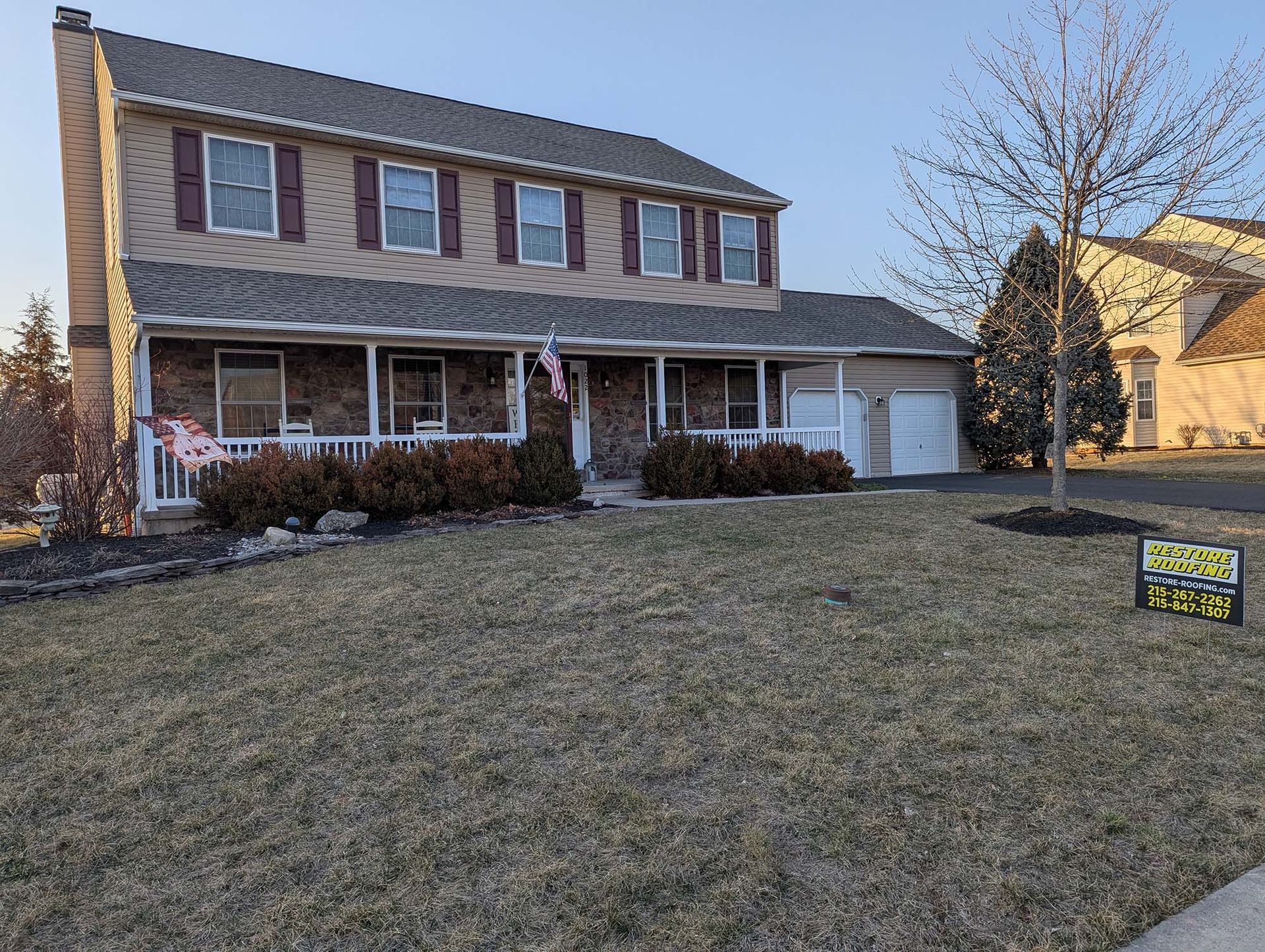 Two-story house with tan siding, brick chimney, and a porch. The lawn is brown.