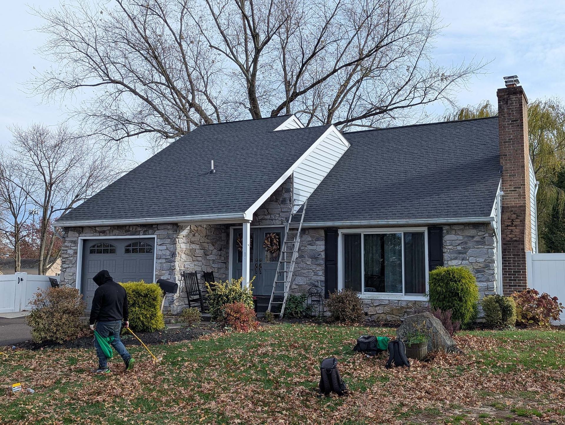 Person raking leaves in front yard of a house with a dark roof and a chimney.