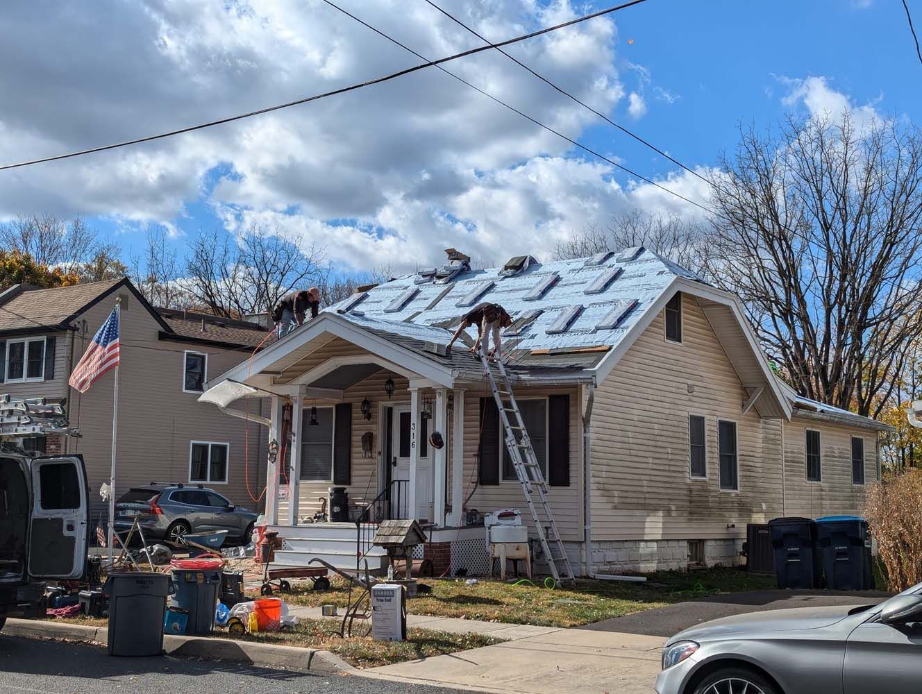 Roofers working on a house with a ladder, blue sky and clouds overhead. An American flag flies next door.