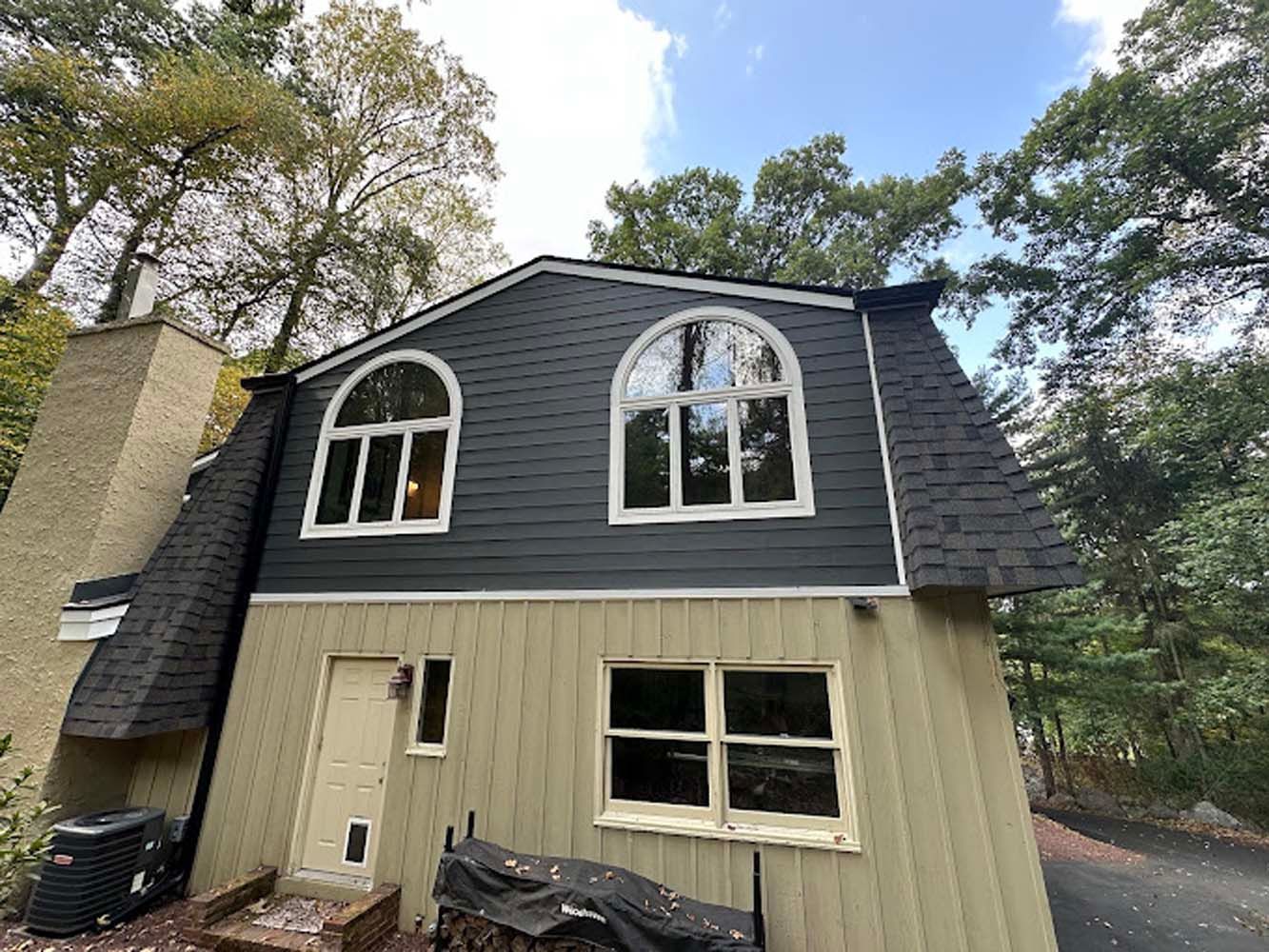 Two-story house with dark gray siding, arched windows, and a tan lower level surrounded by trees.