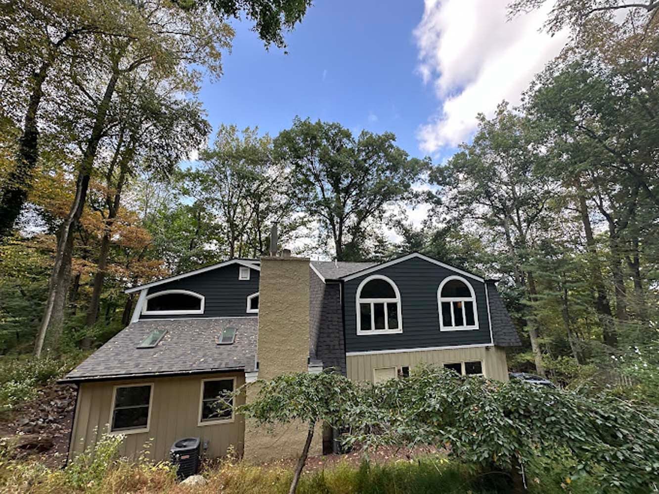 House with dark siding, arched windows, and a tall chimney surrounded by trees under a partly cloudy sky.