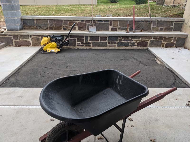 Wheelbarrow in foreground, prepared gravel base for a patio, stone wall and steps in the background.