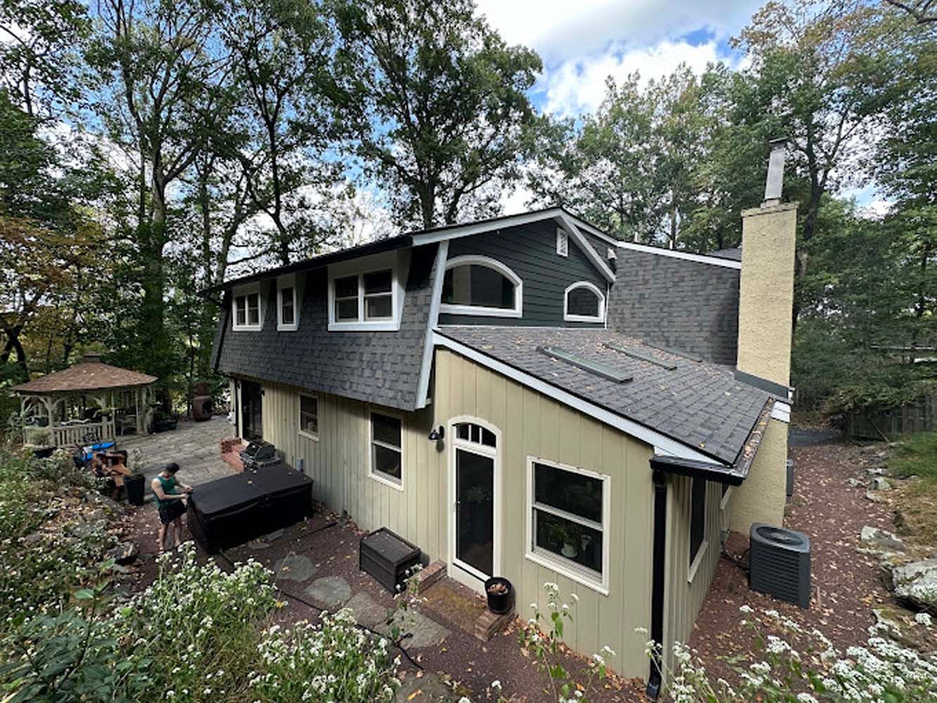 House with gray roof, tan siding, and black hot tub surrounded by trees and a gazebo.