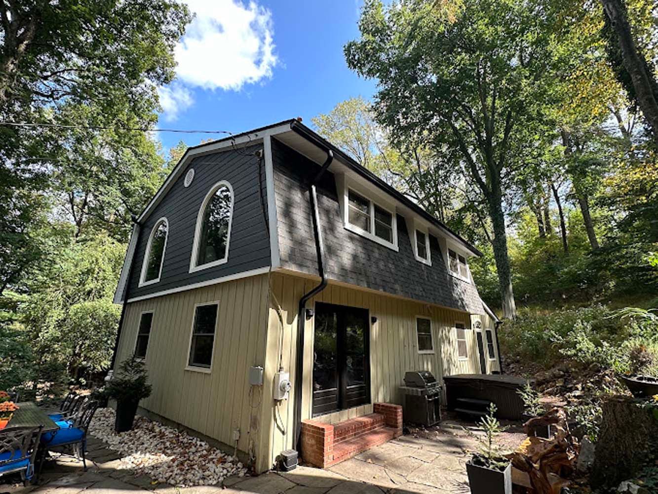 Two-story house with gray roof and light yellow siding, surrounded by trees. Blue sky visible.