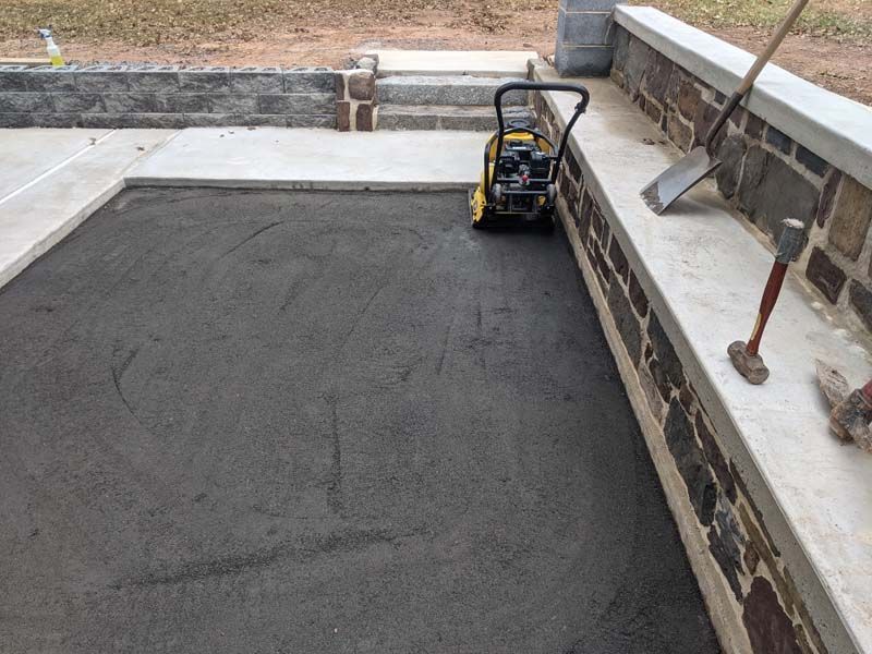 A compact asphalt area is being compacted with a vibrating plate compactor, alongside a stone wall with tools.
