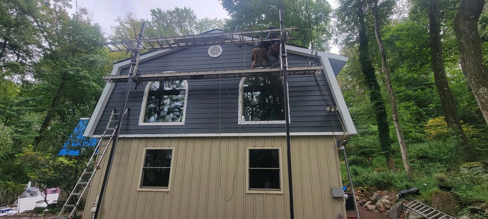 House exterior under construction, surrounded by trees. Scaffolding, ladder, and a partially shingled roof.