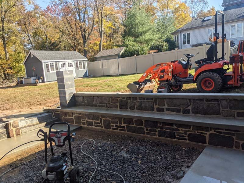 A construction scene: stone wall being built with a tractor, shed, and pressure washer in a yard.