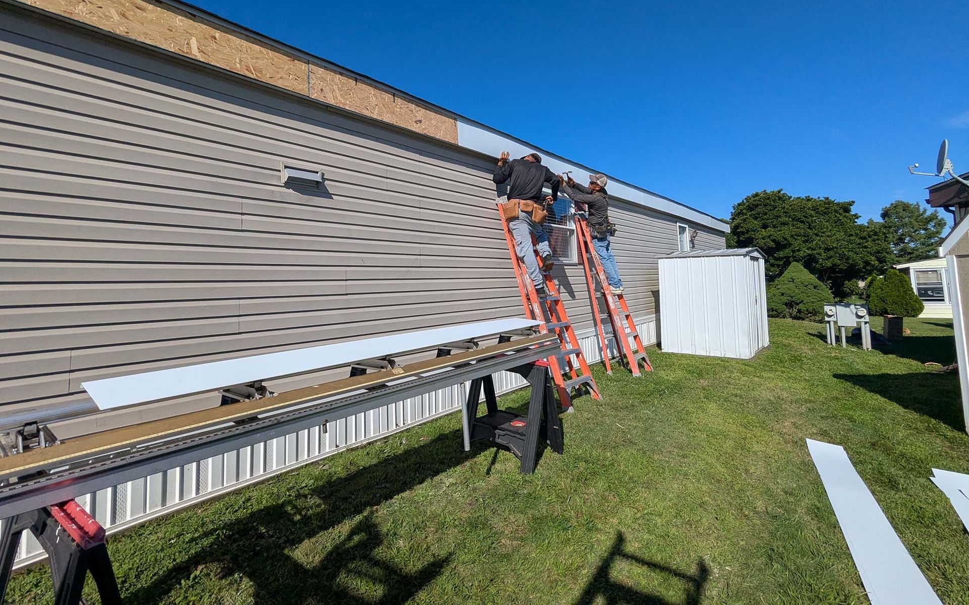 Two people on ladders siding a light brown building; a cutting table with material is set up on the lawn.