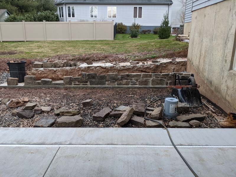 Construction site with stone retaining wall being built next to a building and lawn.