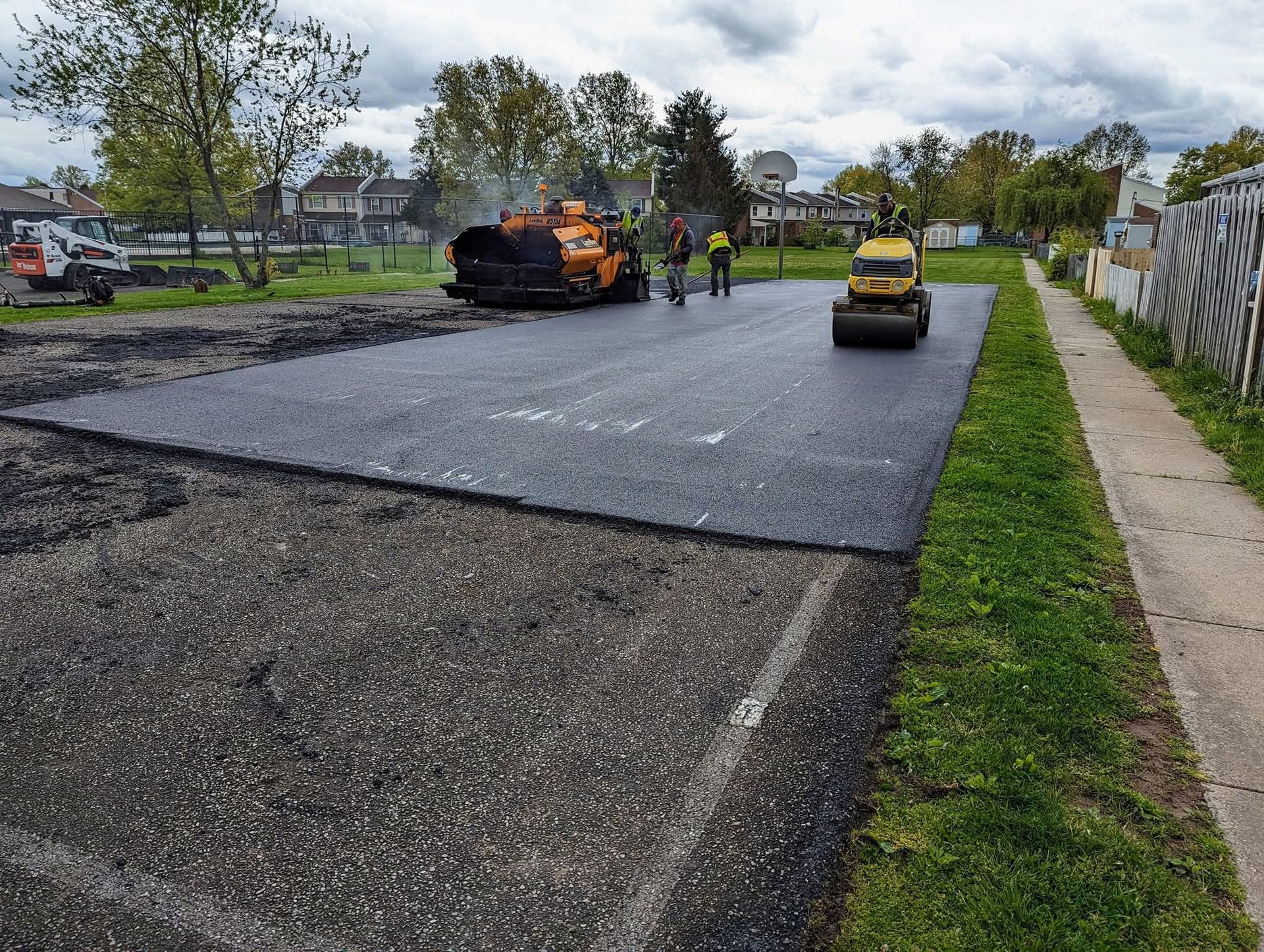 Asphalt paving in progress: machine laying fresh asphalt, workers nearby, roller compacting. Green grass on edge.