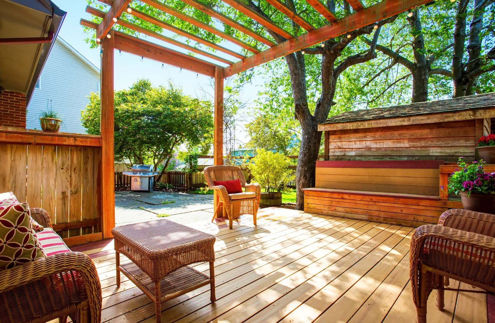 Wooden deck with wicker furniture under a pergola, lush greenery in the background.