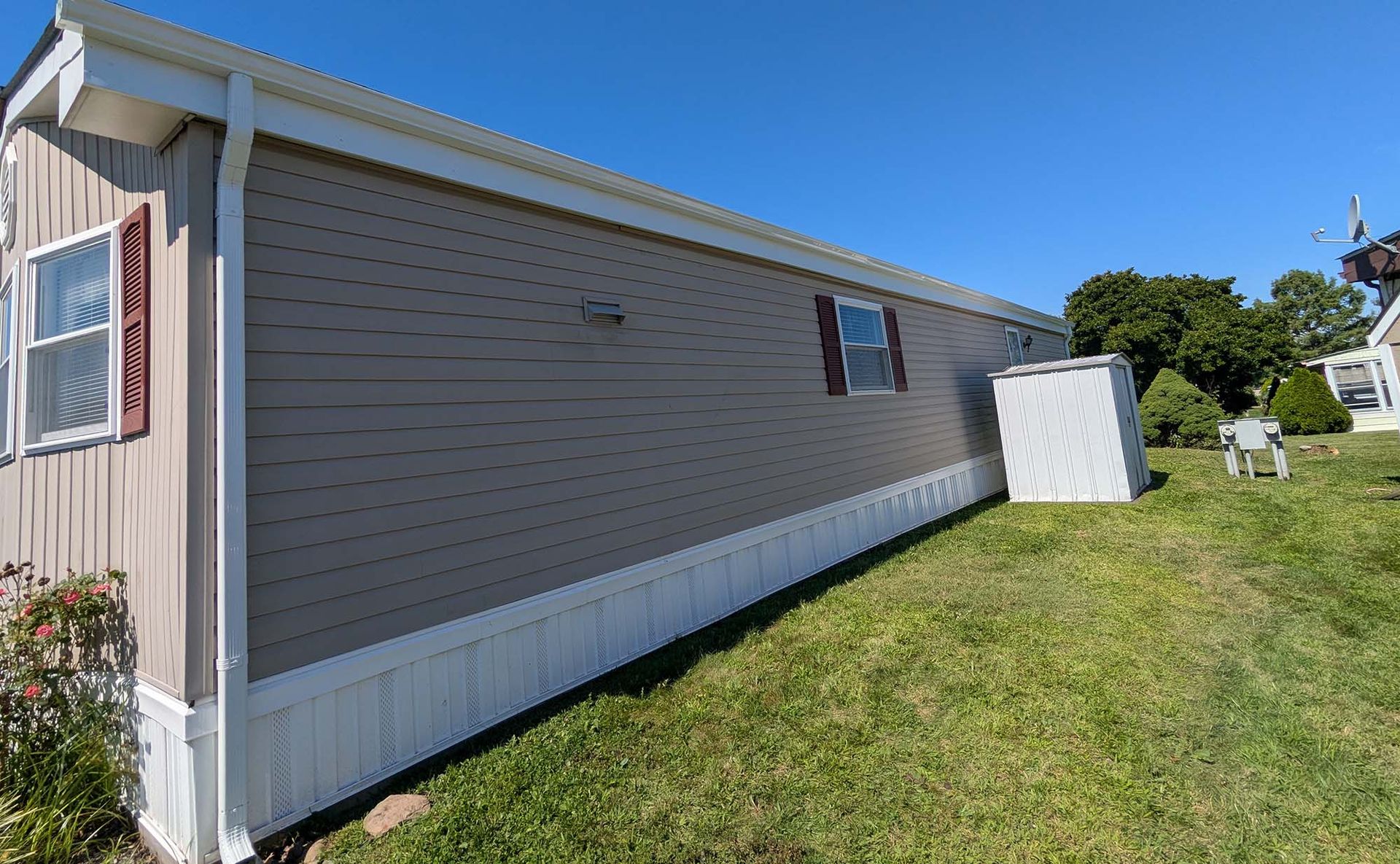 Mobile home with brown siding and white trim on a grassy hill under a blue sky.