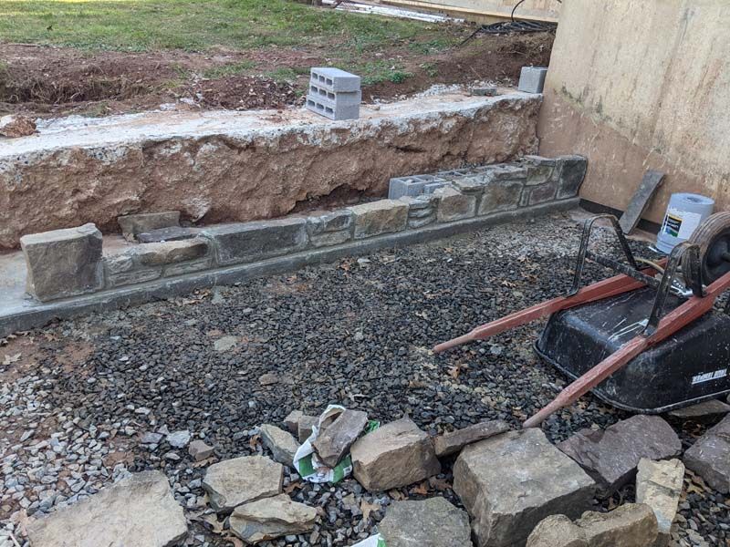 Construction site with a partially built stone retaining wall; dirt, gravel, tools visible.