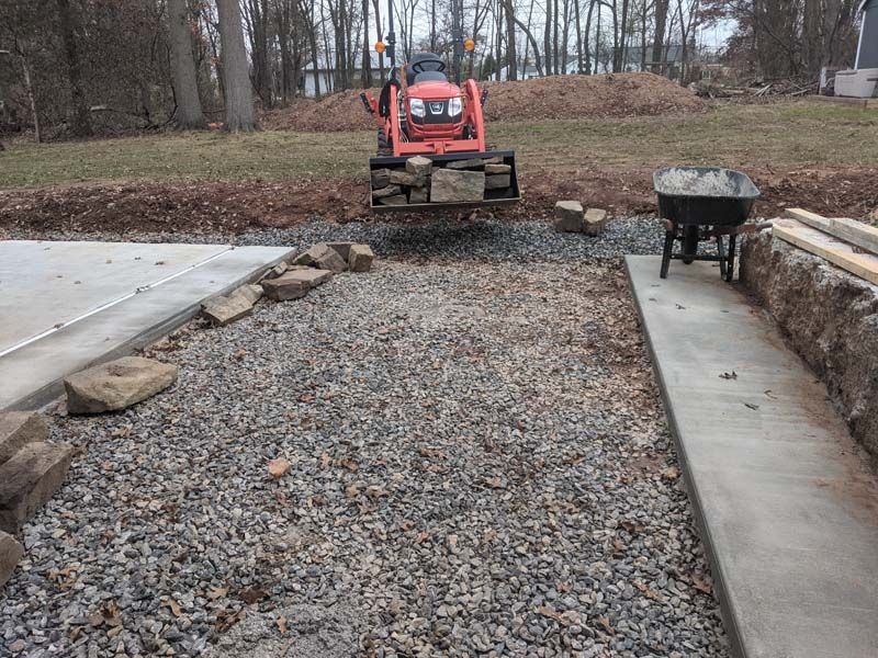 Tractor loading rocks into a gravel area near a concrete border. Wheelbarrow, wood beams, and trees are visible.
