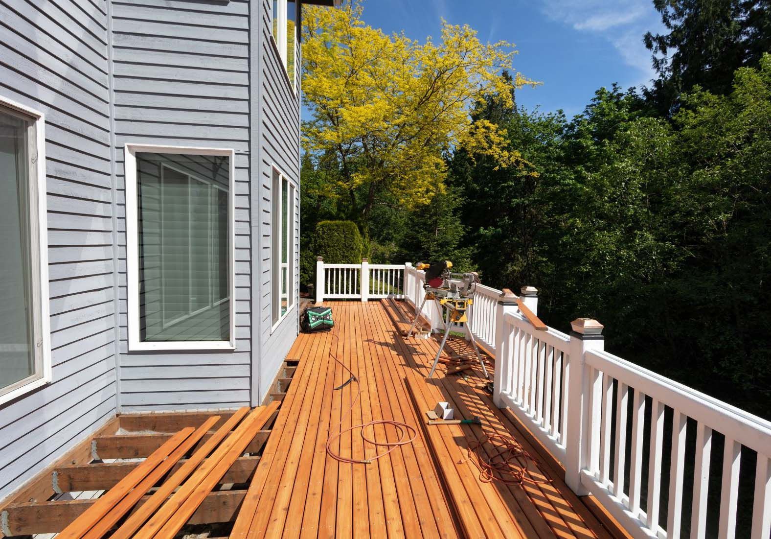 Deck construction with exposed wood beams, new wood planks, and white railing next to a gray house.
