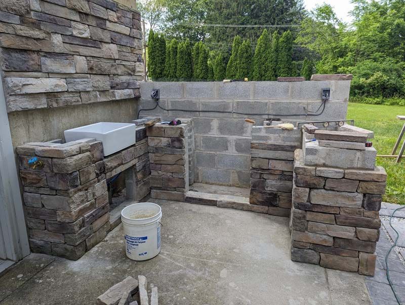 Stone masonry outdoor kitchen under construction. Gray stone, concrete blocks, and a white sink are visible.
