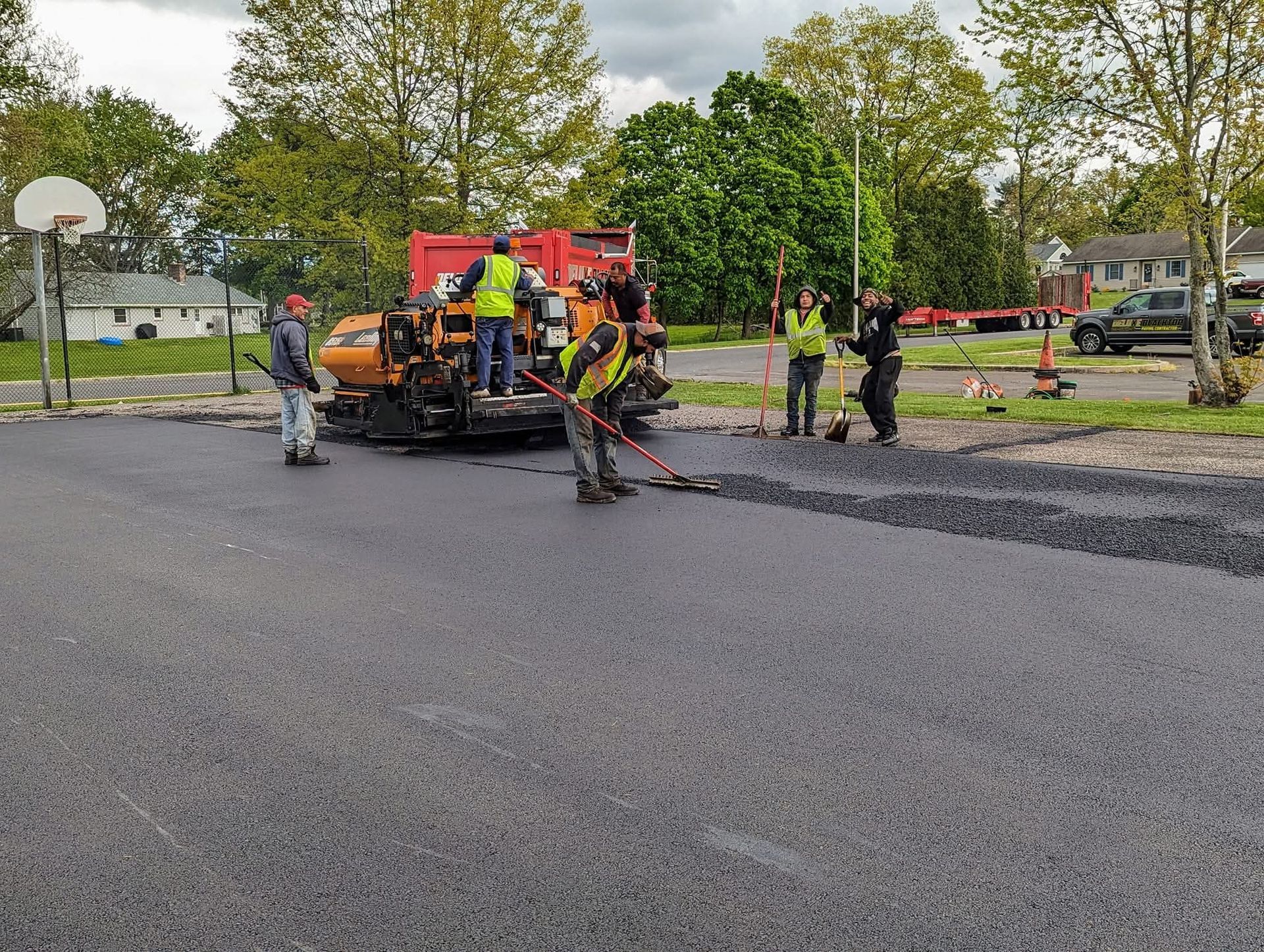 Asphalt paving crew working on a basketball court. Dark gray asphalt, machinery, workers with tools, trees in the background.