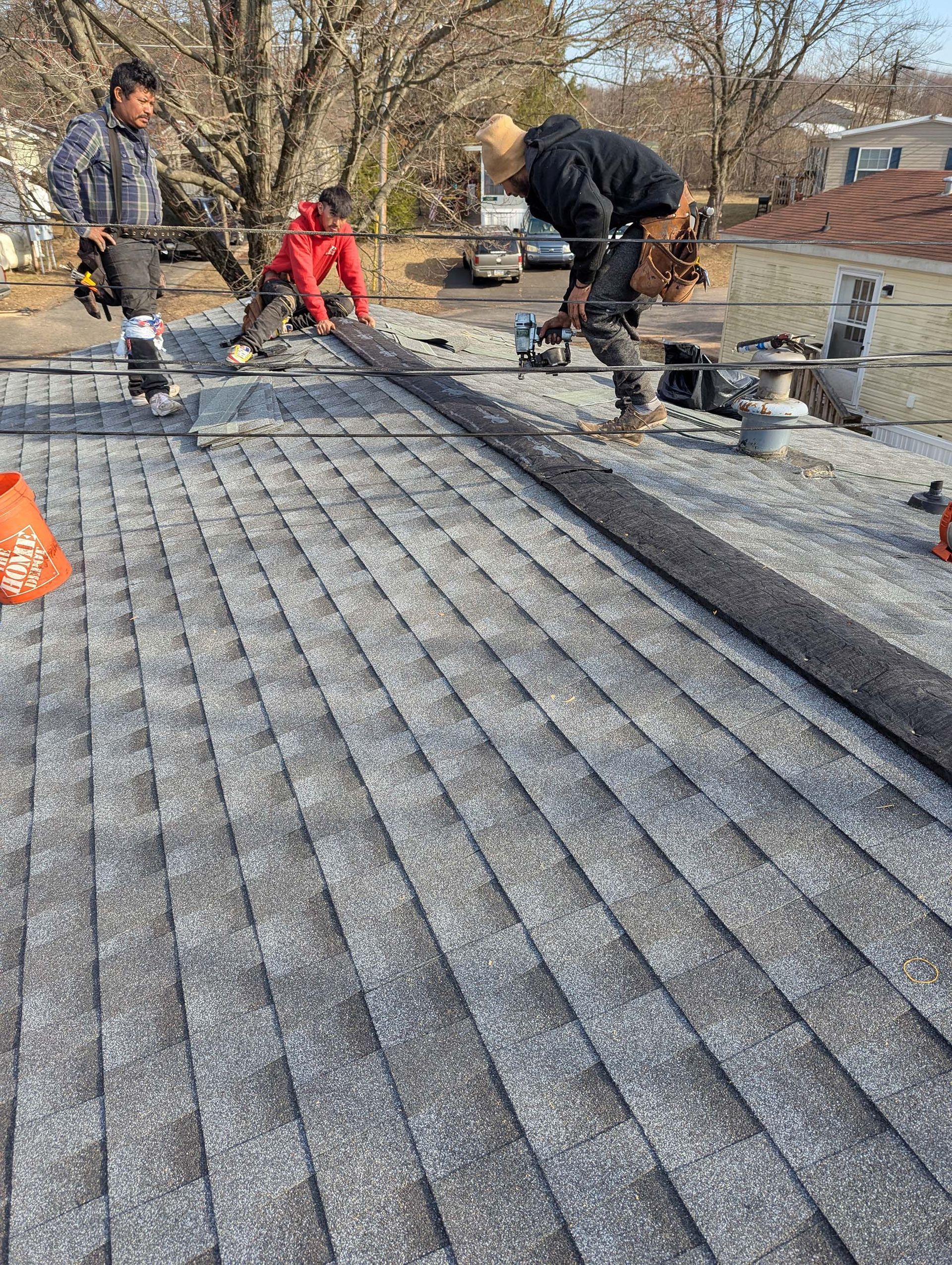 Roofers installing roofing material. One lays strip, two work with tools on a rooftop.