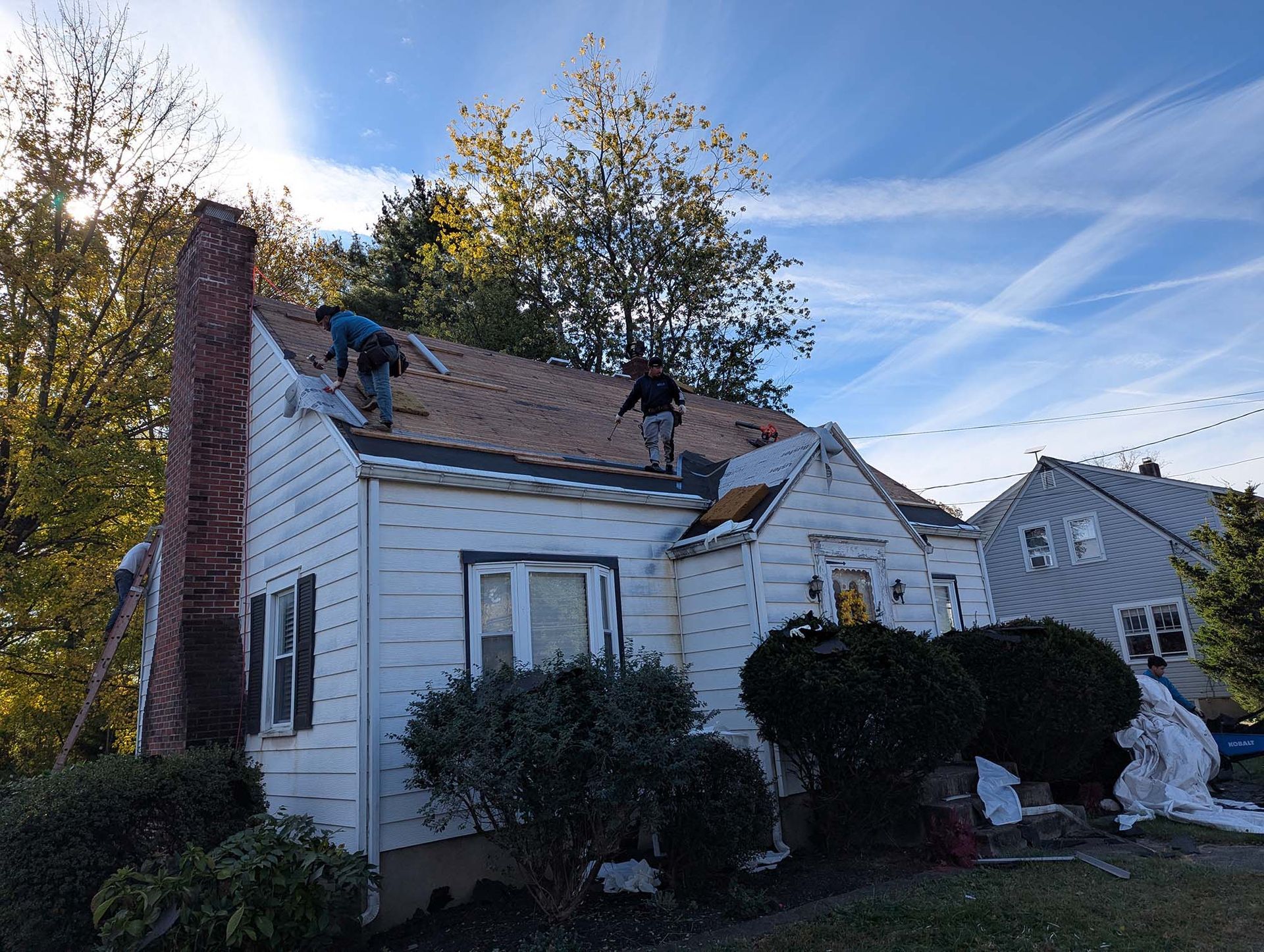 Two roofers working on a house roof on a sunny day. A brick chimney is on the left.