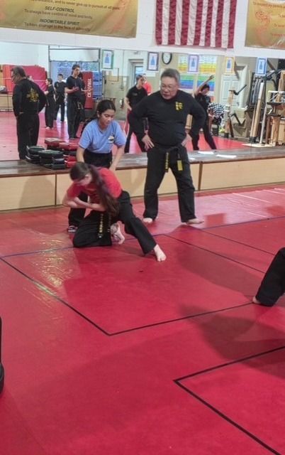 Martial arts training in a red-matted dojo, with two people practicing a takedown and an instructor observing.
