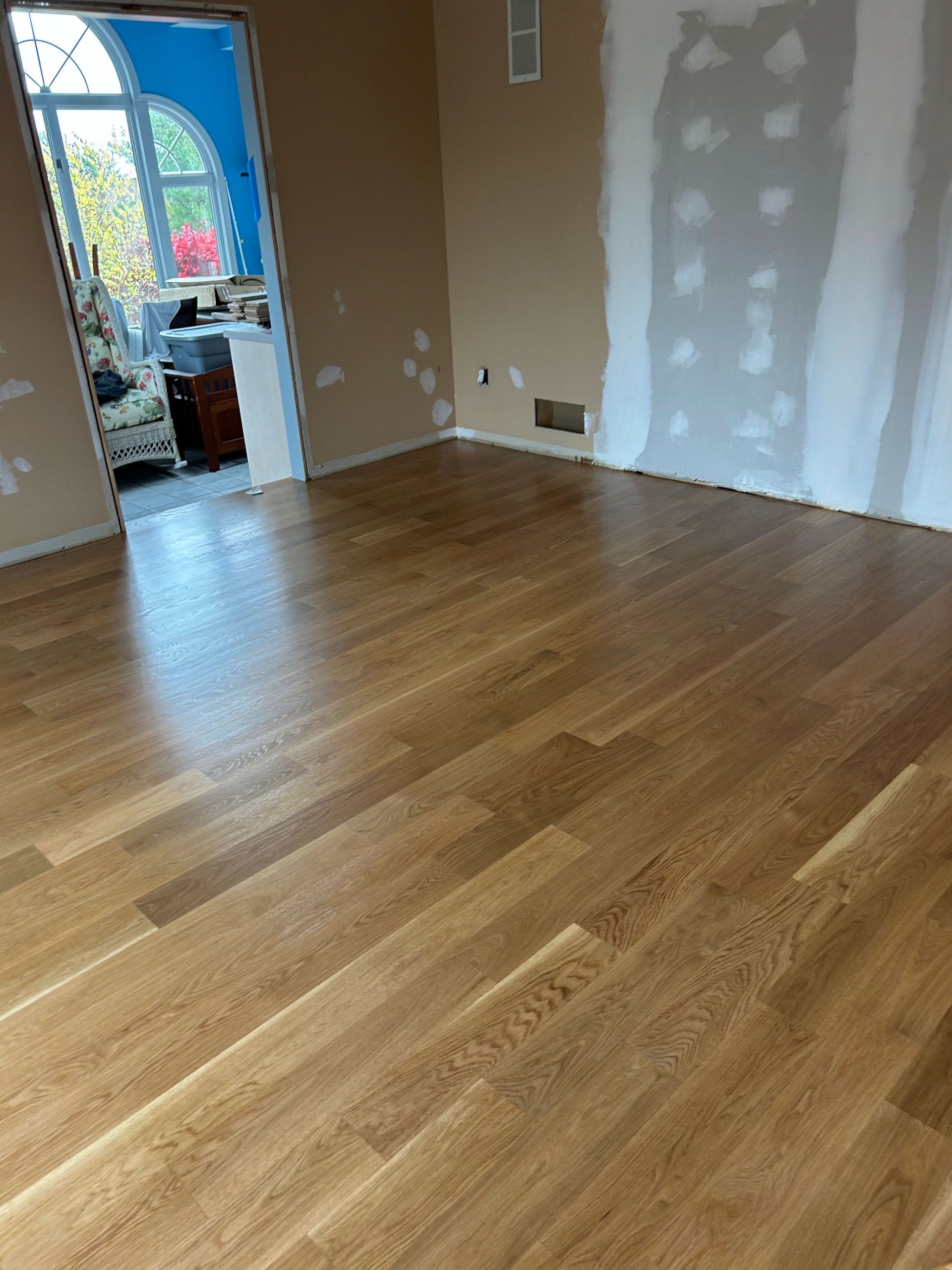 Empty room with hardwood floor, unfinished drywall patches, and an open doorway to a bright adjoining space