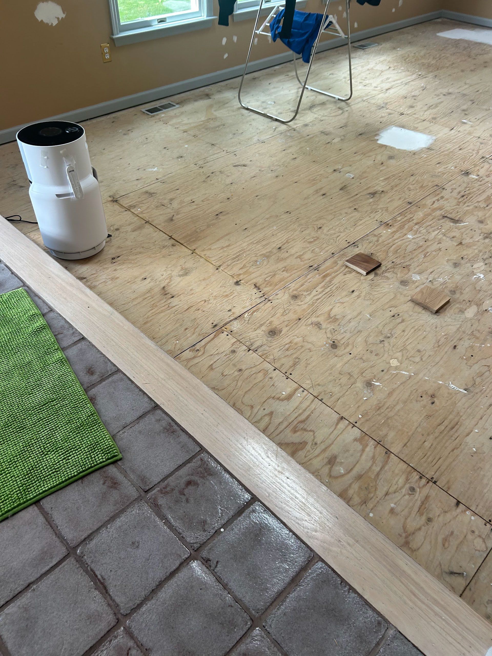 Partially stripped floor with exposed plywood, scattered debris, and a white canister near a windowed room