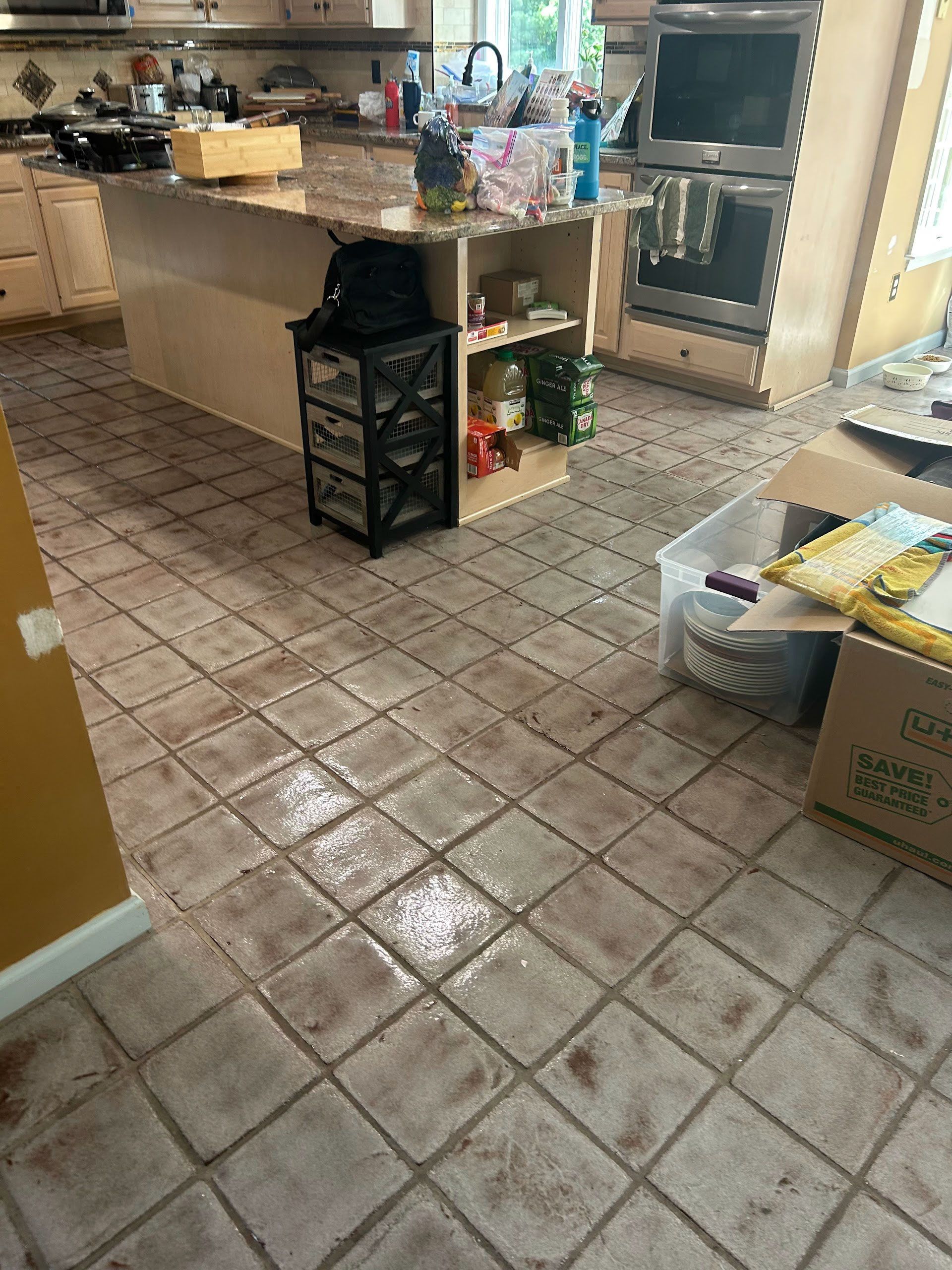 Kitchen with tiled floor, island counter, and a shelving unit beside boxes and cleaning supplies.