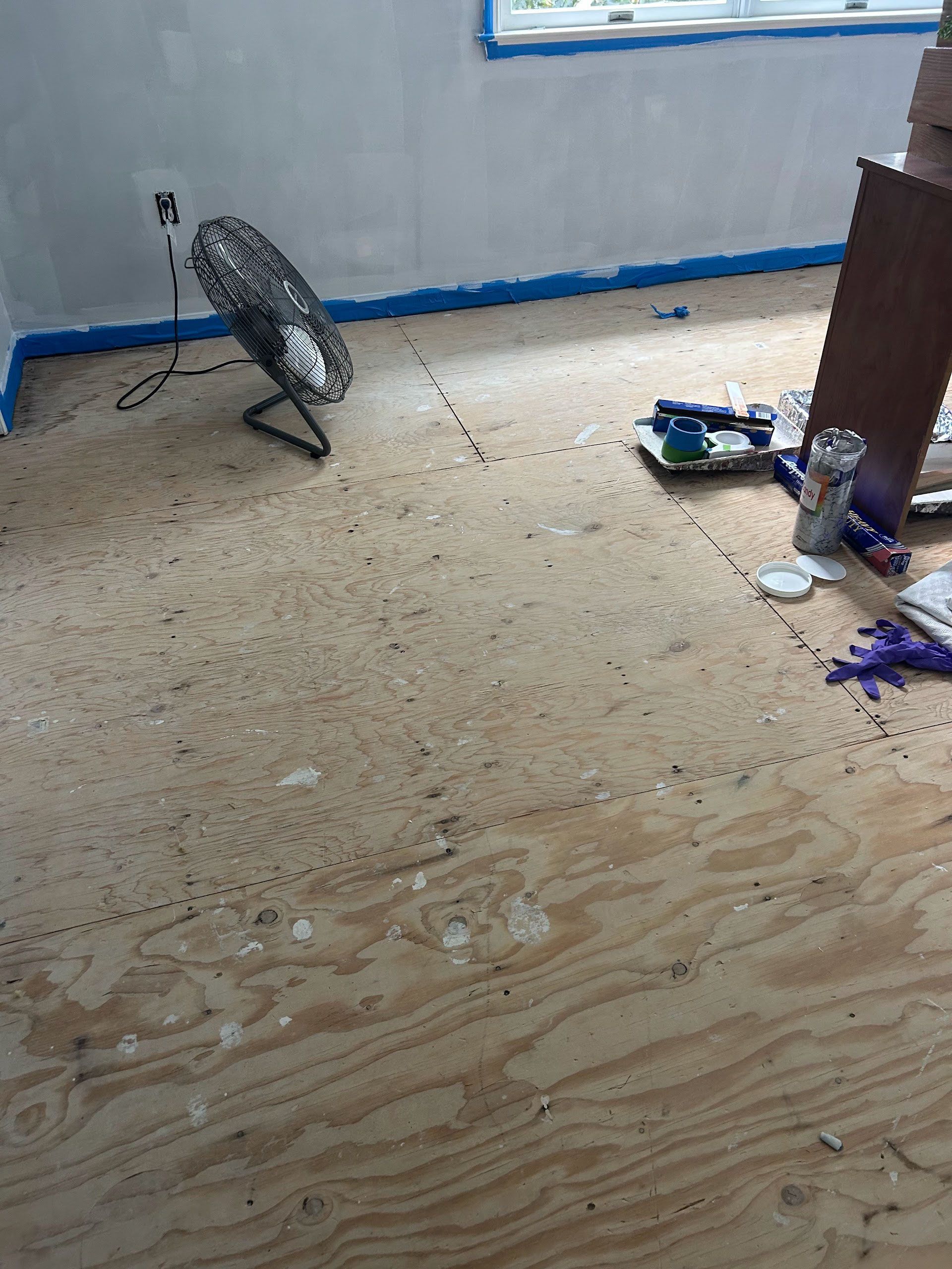 Spilled purple paint on a speckled floor with a standing fan and small tools near a wall.