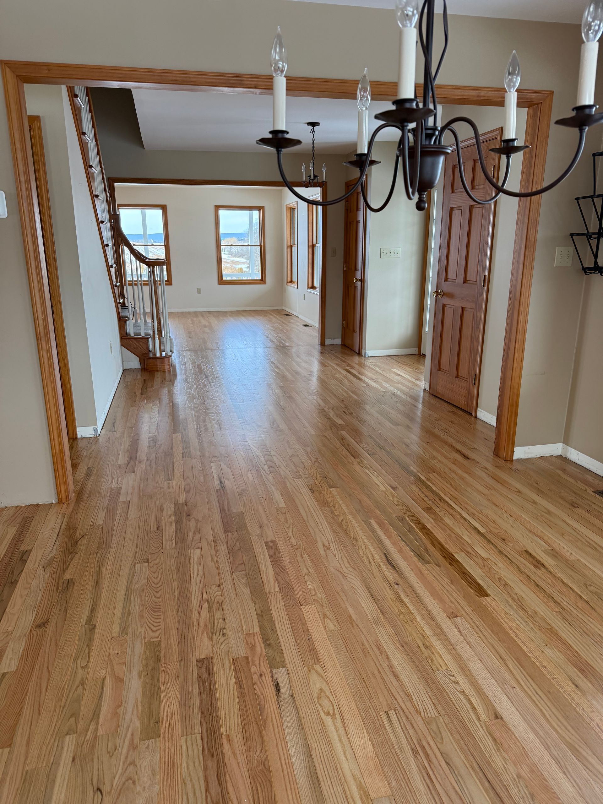 Empty hardwood-floored hallway with chandeliers and a bright window at the far end
