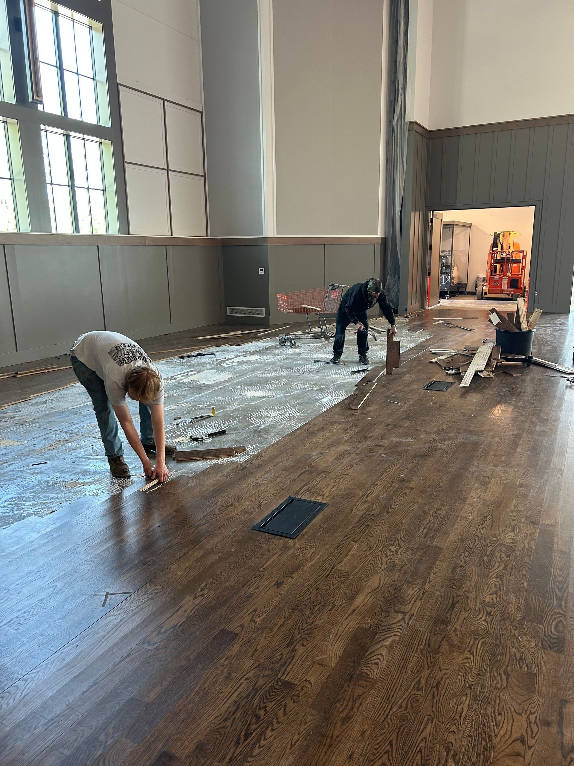 Two people removing flooring in a large room with high ceilings and tall windows.