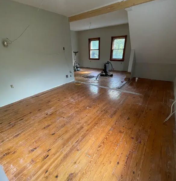 Wooden floor being refinished in a room with light green walls and two windows in the background.