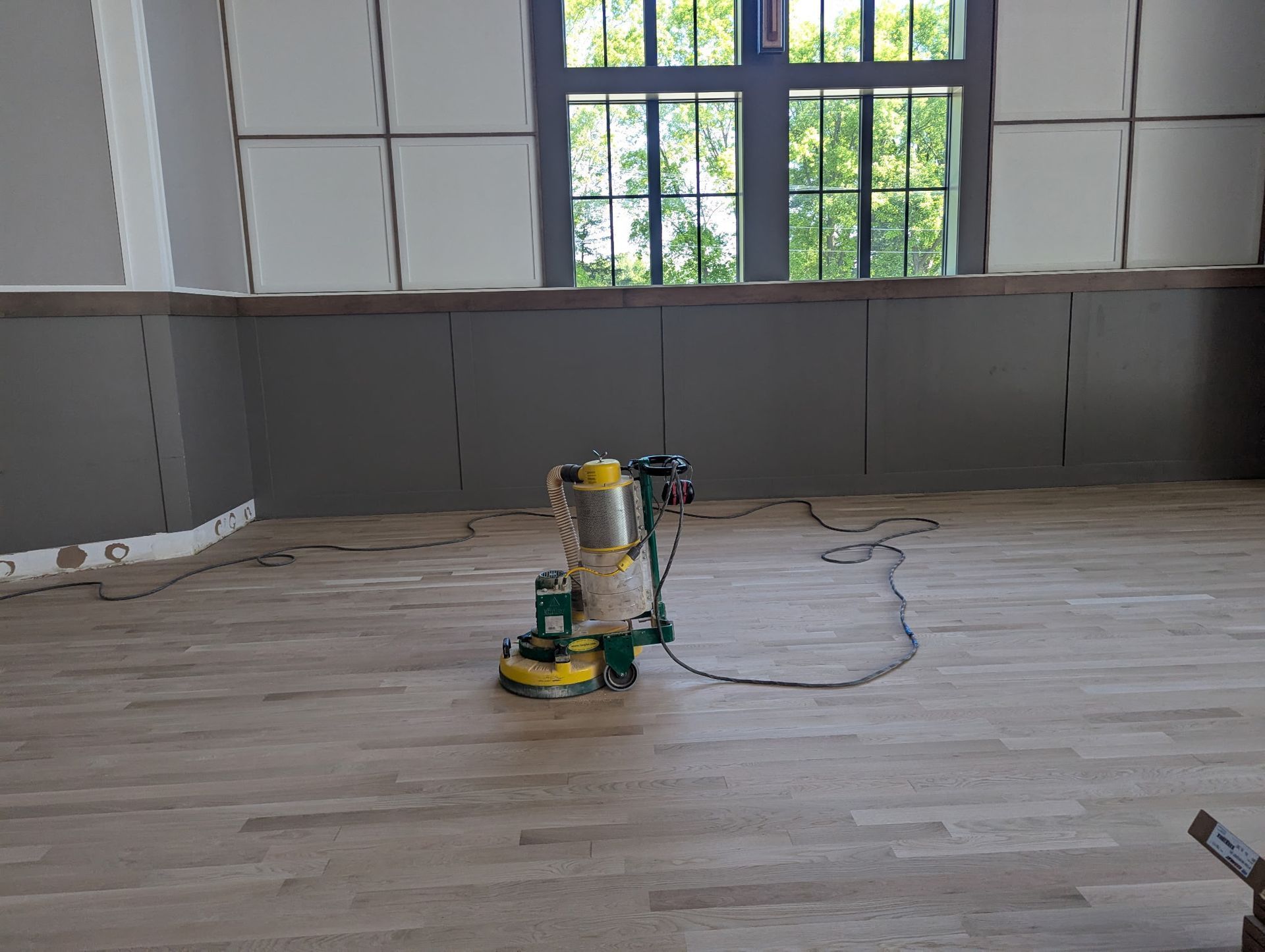 Floor sander on a light wood floor in a room with gray walls, near a window.