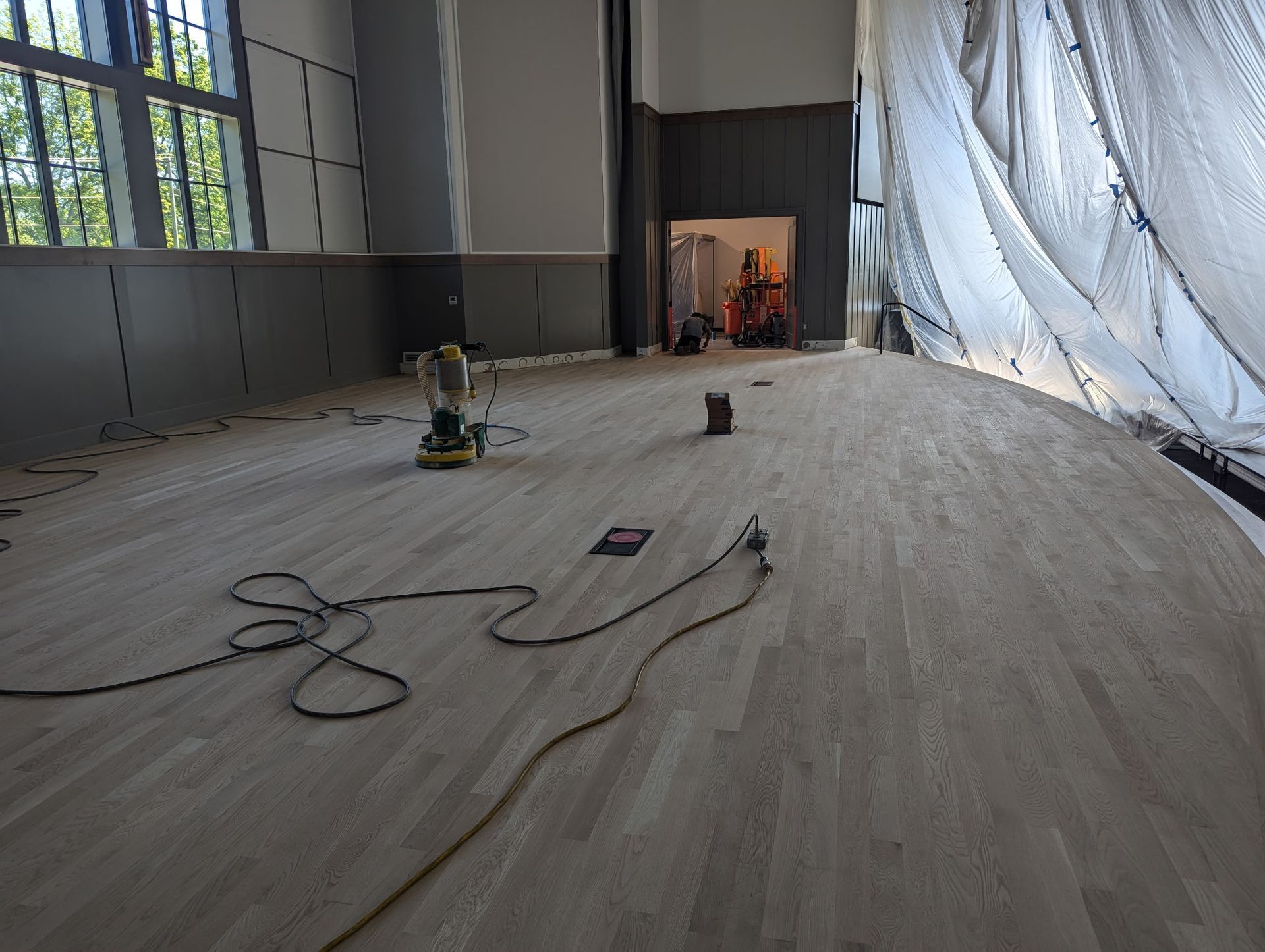 Wooden floor being sanded in a room. A floor sander, cord, and plastic sheet are present.