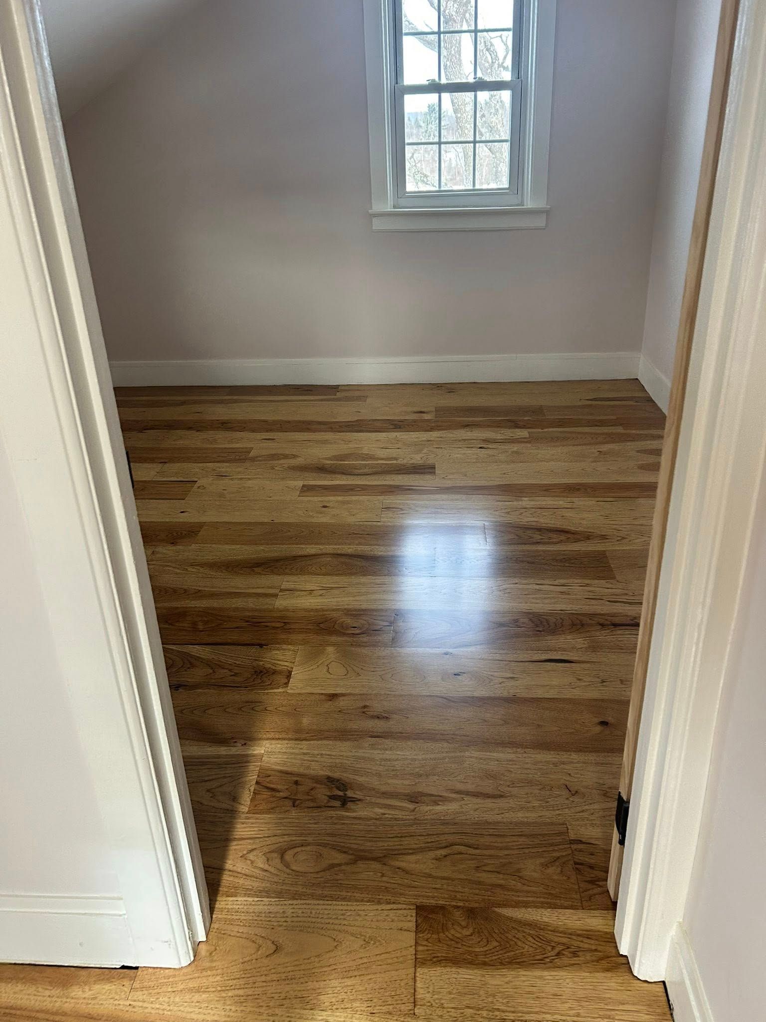Empty small room with polished wood floor, beige walls, and a window letting in daylight.