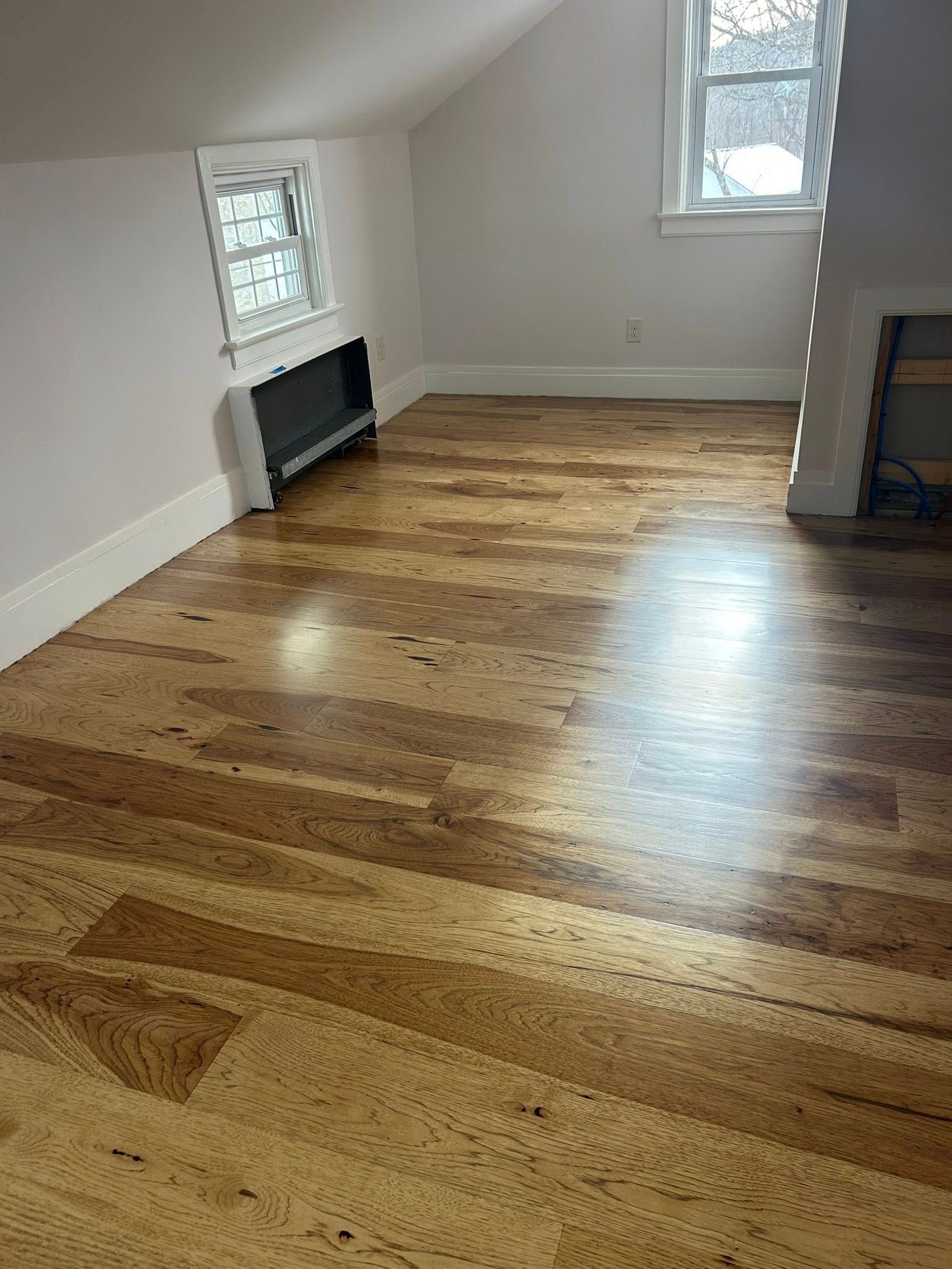 Empty attic room with hardwood floors, sloped ceiling, two windows, and a wall radiator