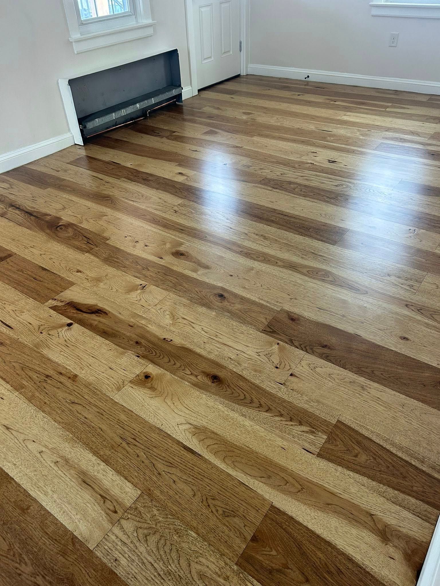 Empty room with polished hardwood floors, white walls, and a wall heater by the door.
