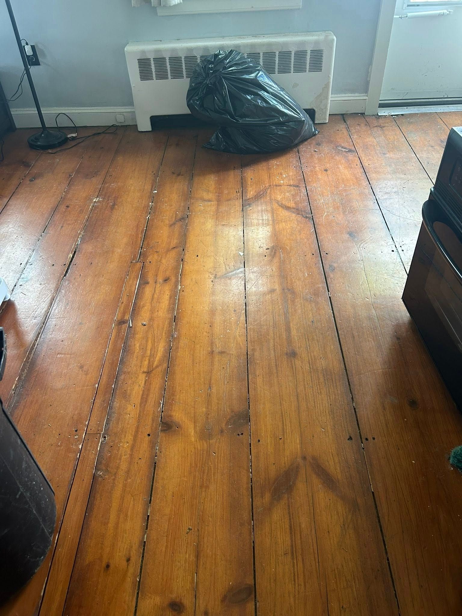 Wooden floor with a black trash bag in front of a wall heater and window light