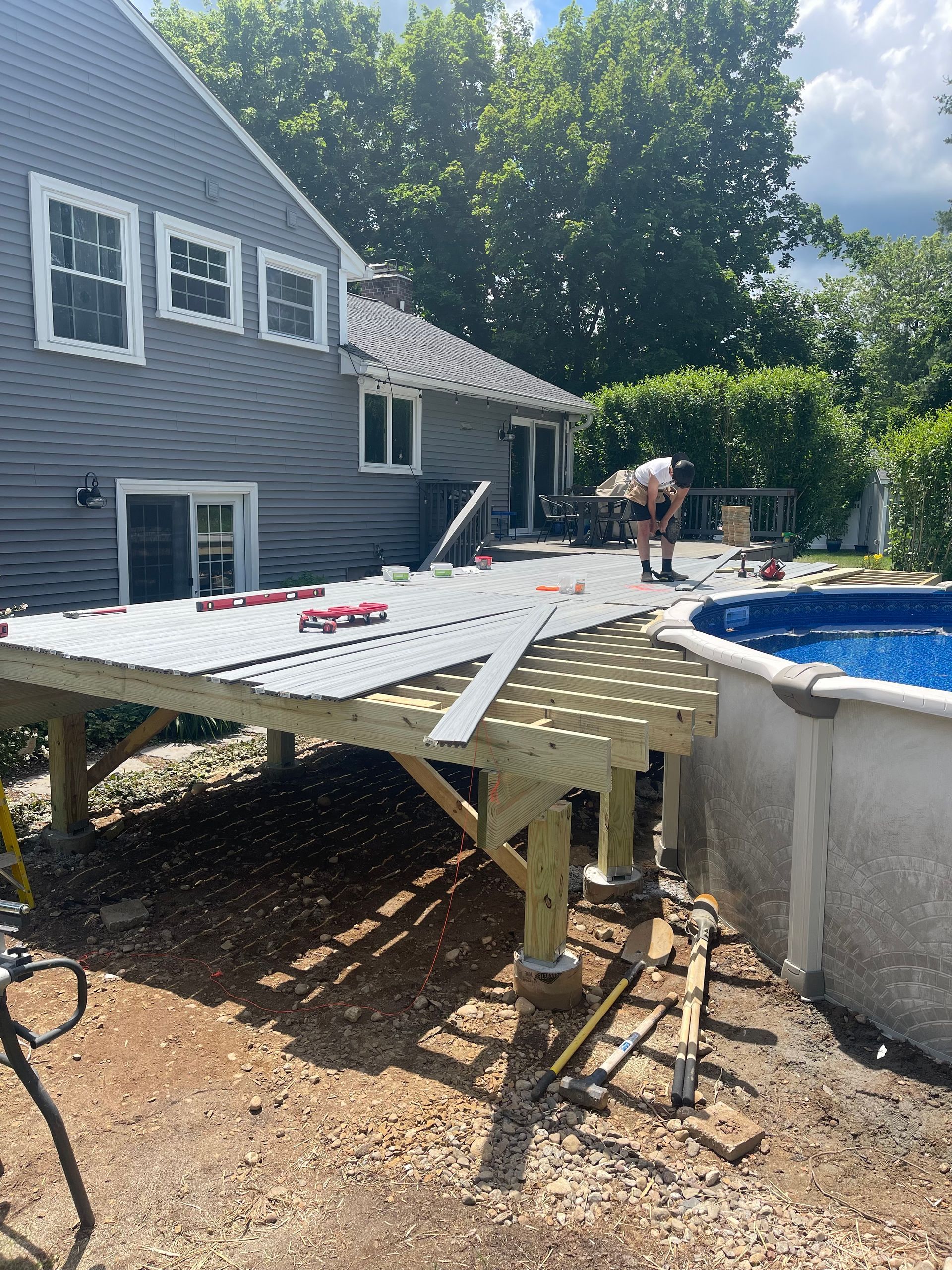 Deck construction near an above-ground pool. A person works on the partially built structure, next to a house on a sunny day.
