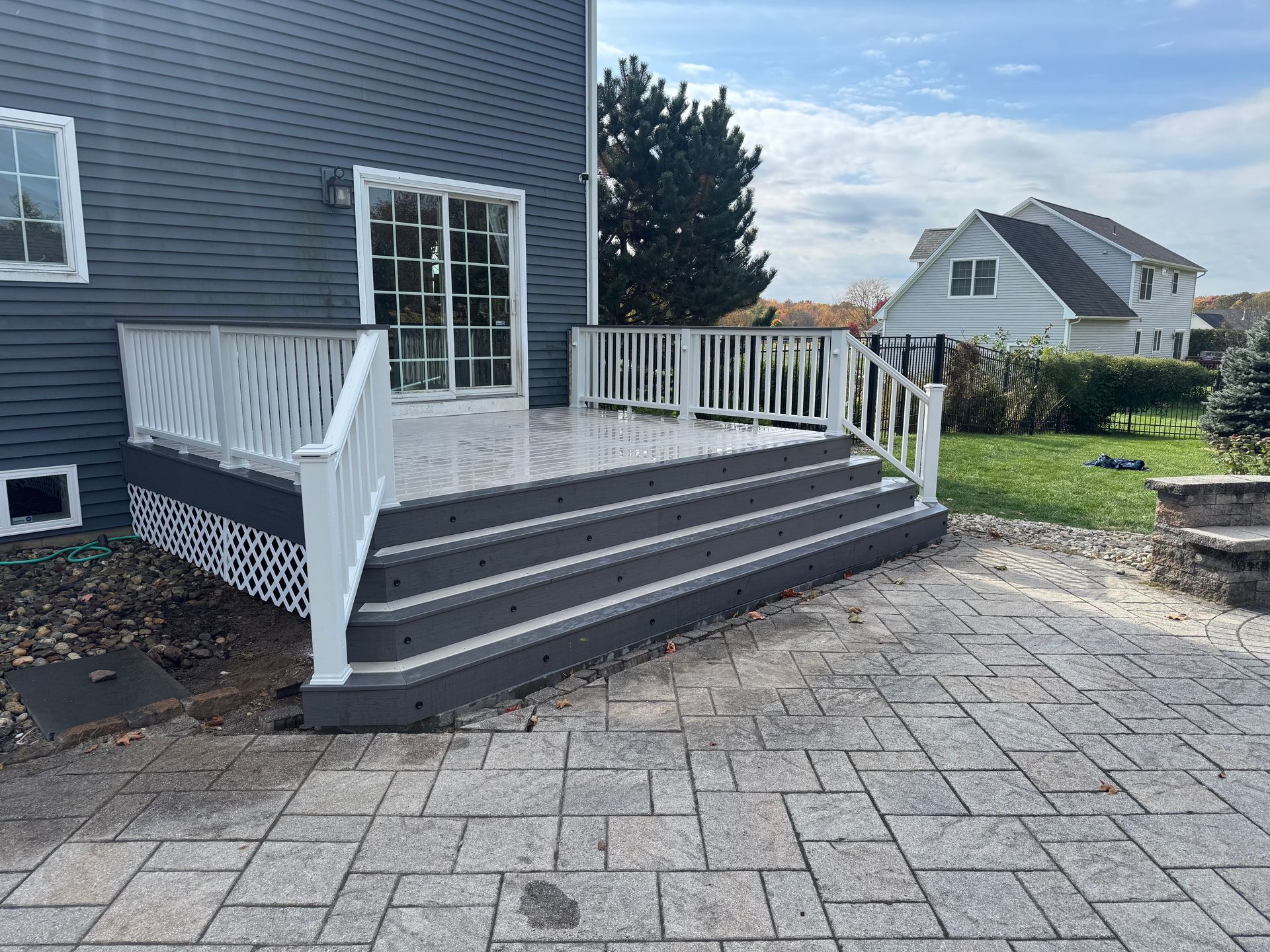 Grey deck with white railing and steps leading to a gray stone patio beside a dark grey house.