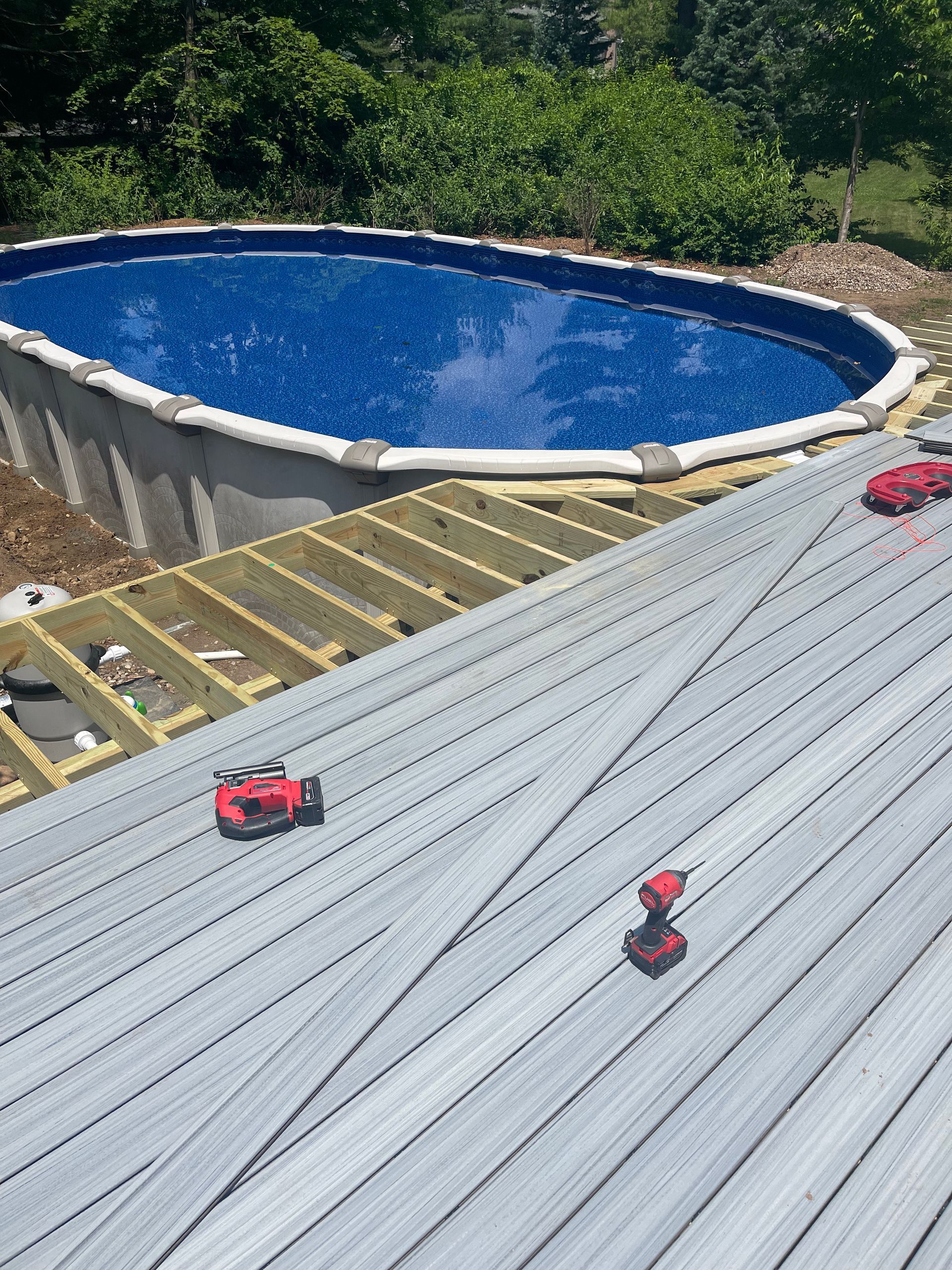 Deck construction near an oval pool; gray planks, red power tools, and blue pool cover.