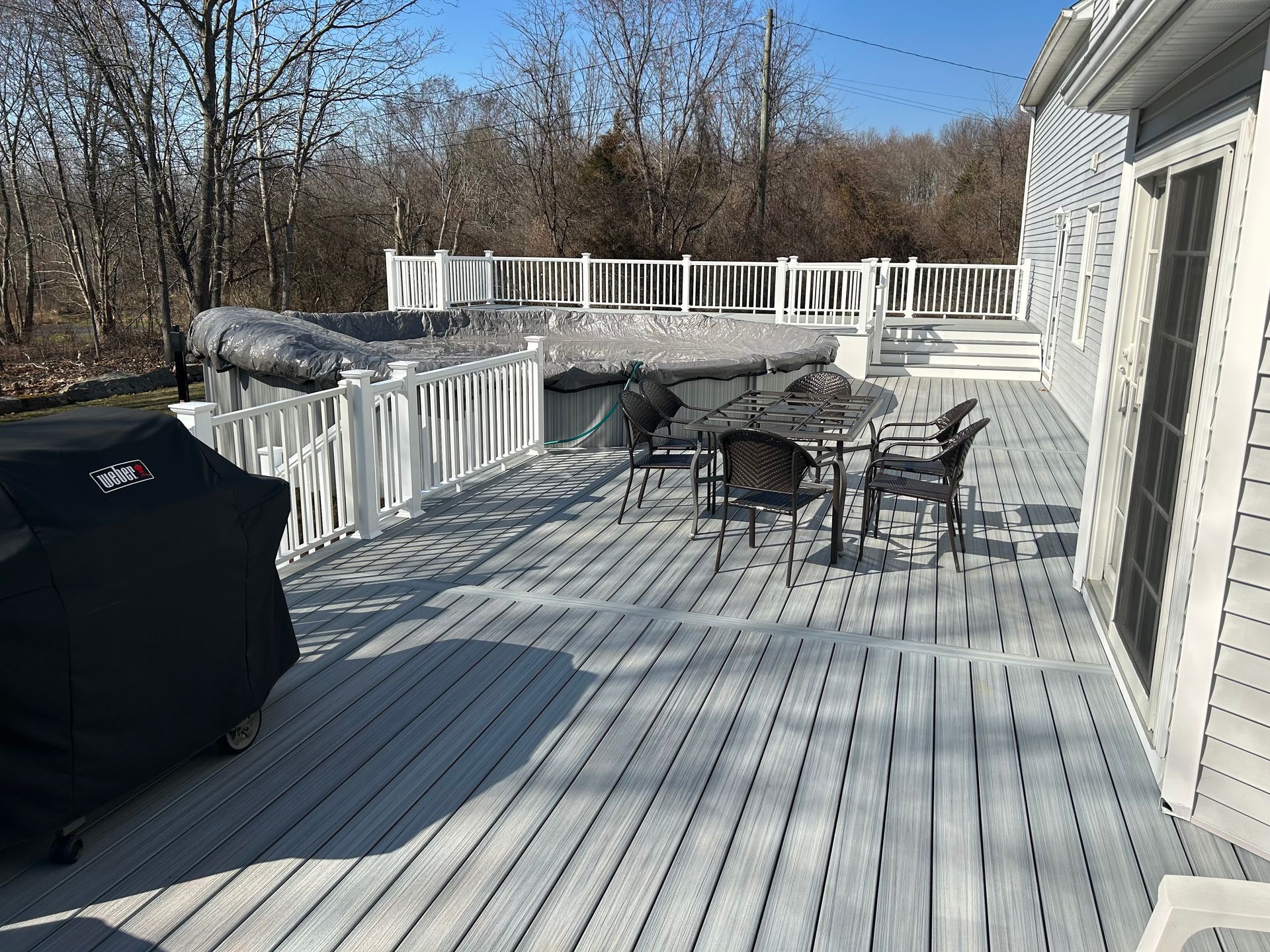 Deck with covered hot tub, table, chairs, and grill next to a house in an outdoor setting.
