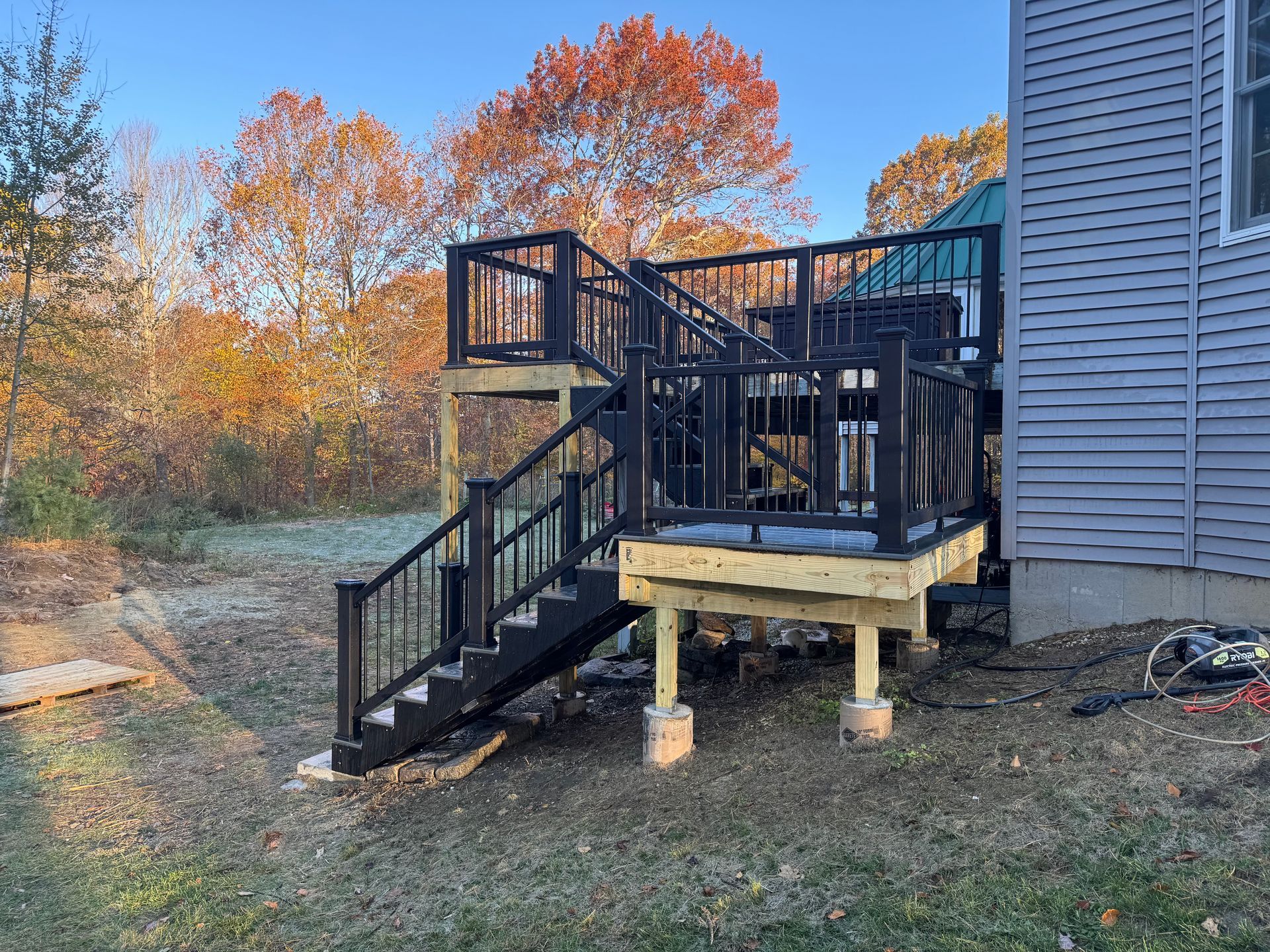 Black deck with stairs attached to a gray house, surrounded by grass and fall foliage.
