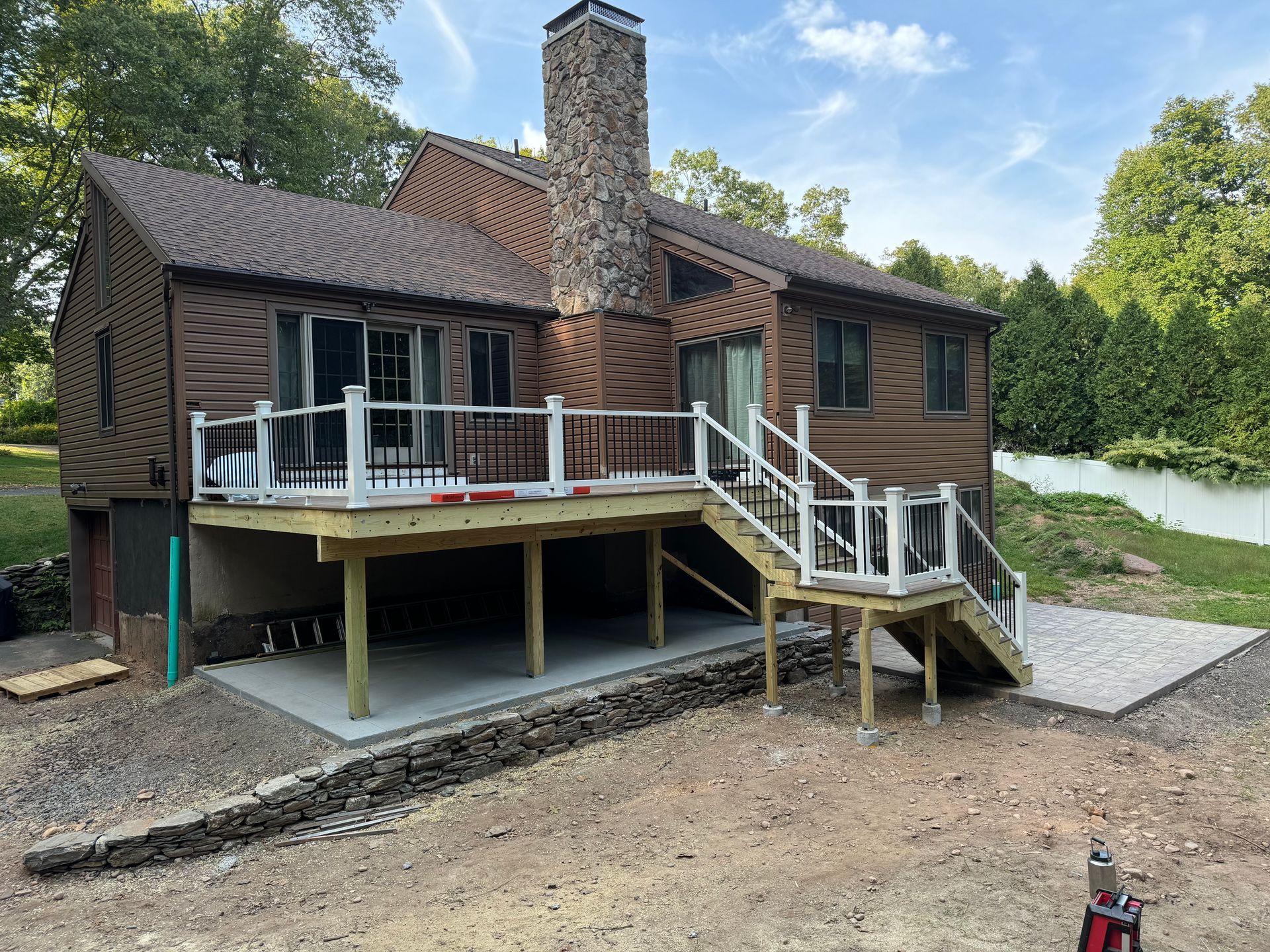Two-story brown house with a new deck and concrete patio, next to a gravel yard, under a blue sky.