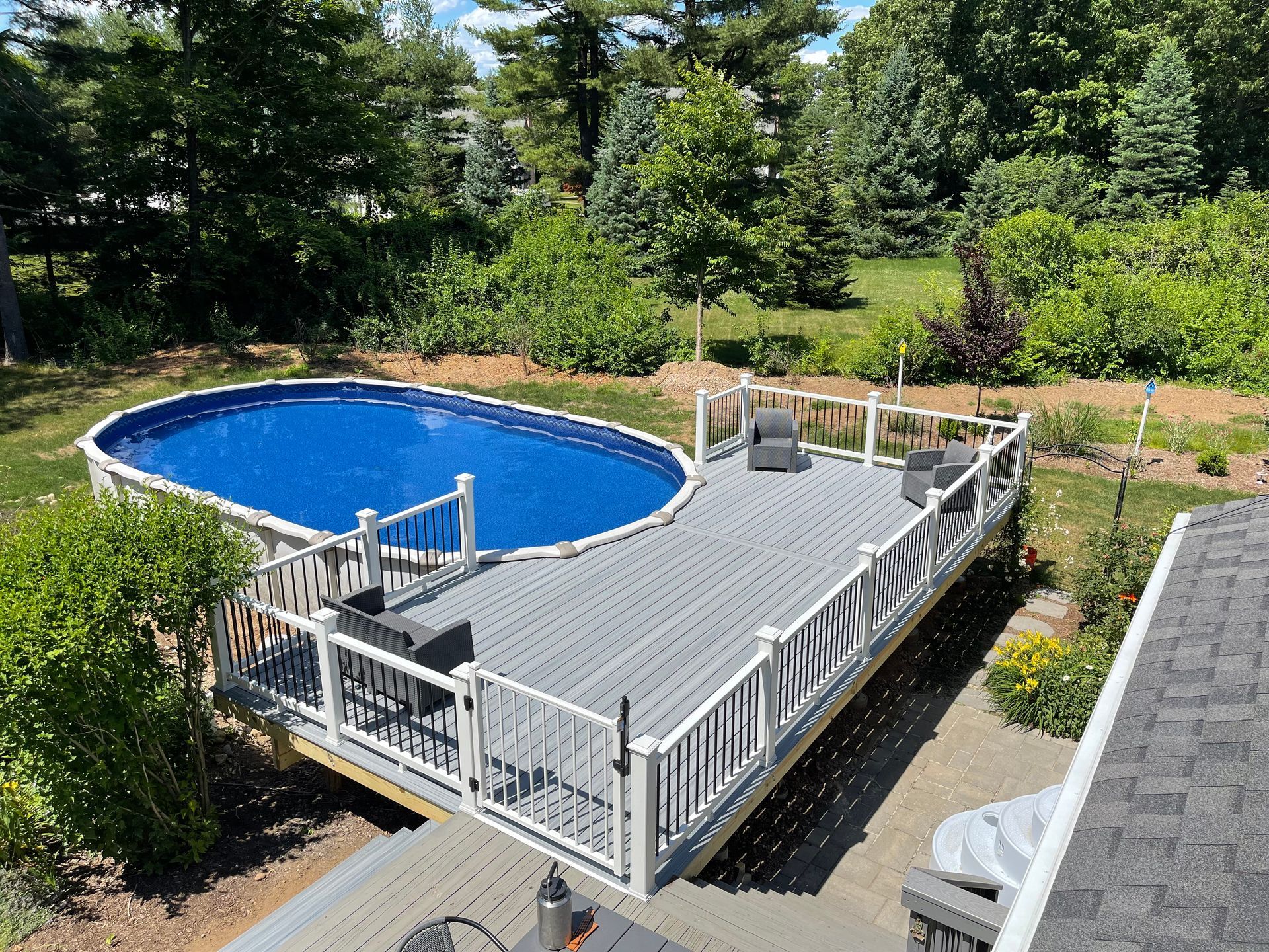 Above-ground oval pool with blue cover and gray deck with seating, in a backyard.
