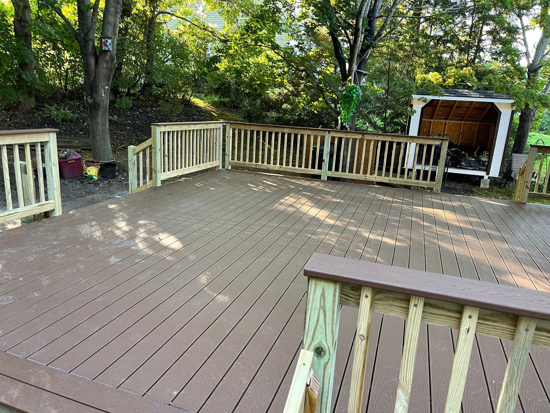 Wooden deck with railing, leading to a forested area and a firewood shed. Brown deck, light wood railing.