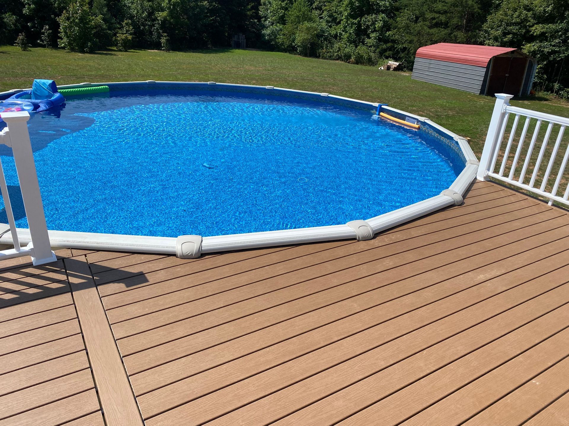 Above-ground pool surrounded by a brown composite deck and white railing. Lush green lawn and trees in the background.