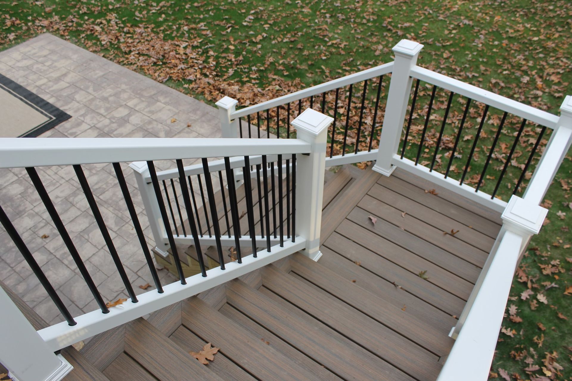 White deck railing with black vertical bars, leading down composite stairs, overlooking a patio and lawn.