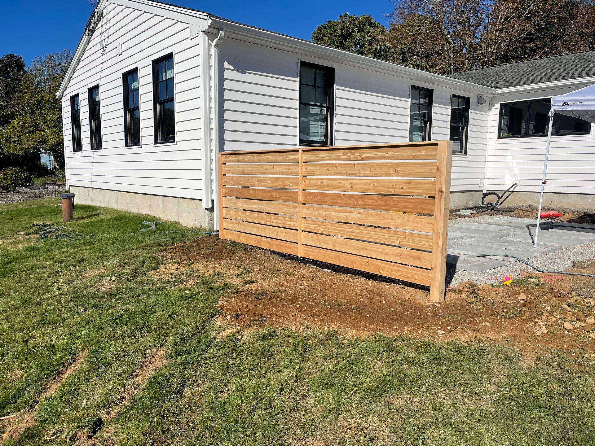 Wooden fence next to a white house with black window frames and a partially completed patio.