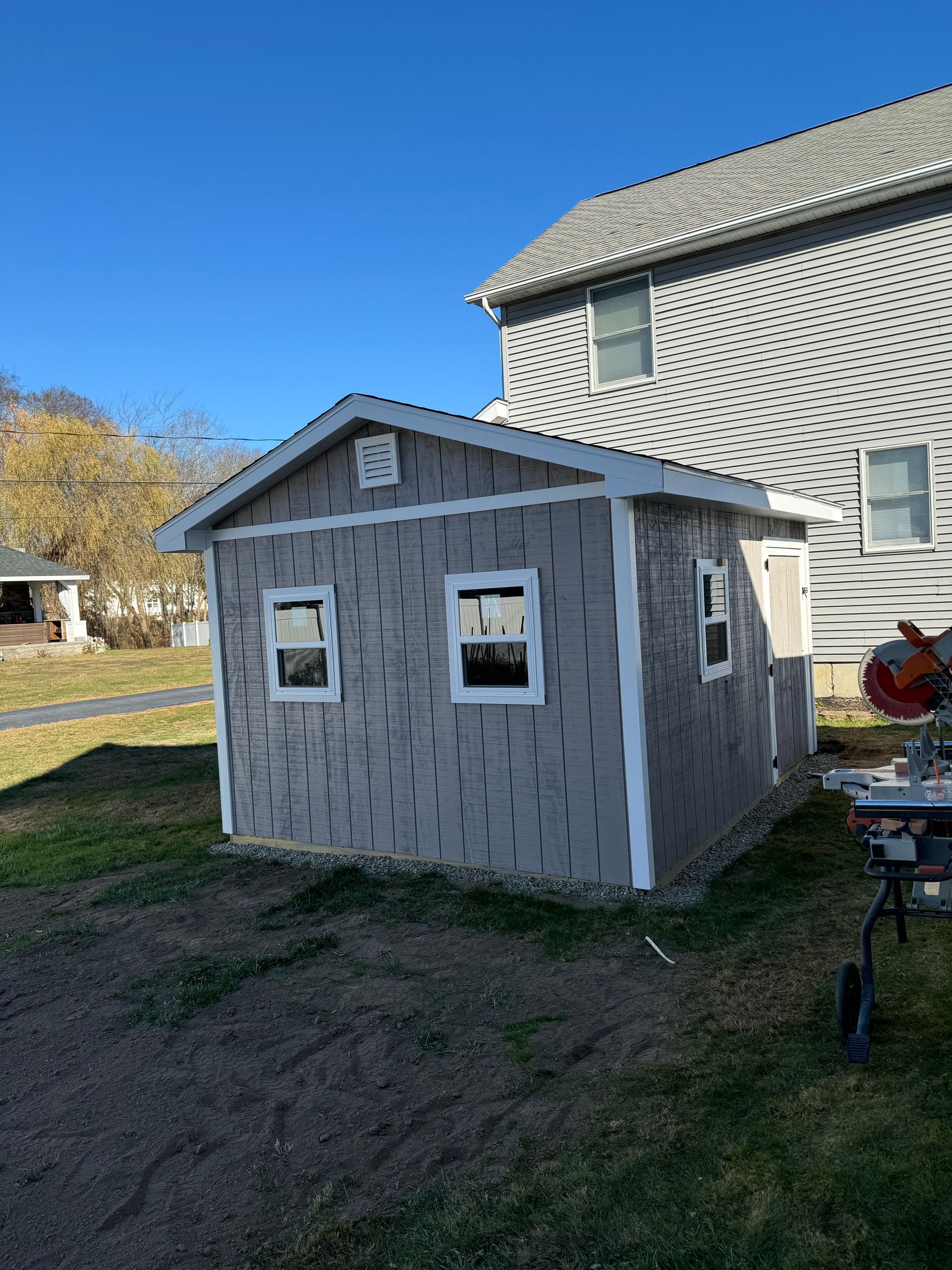 Gray shed with white trim, two windows, next to a two-story house under a blue sky.