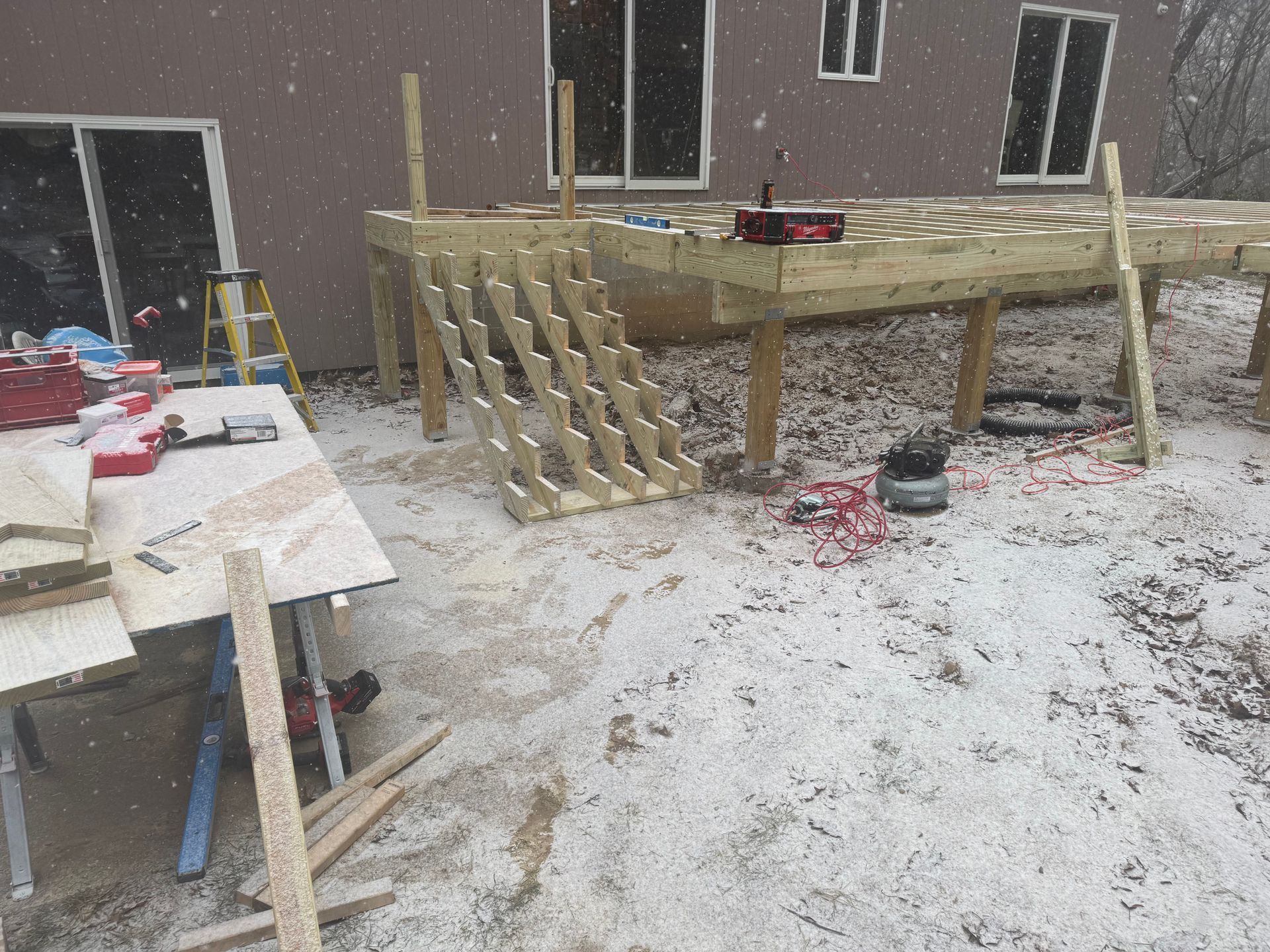 Deck construction in snowy conditions. Stairs and wooden platform are visible, surrounded by snow and tools.